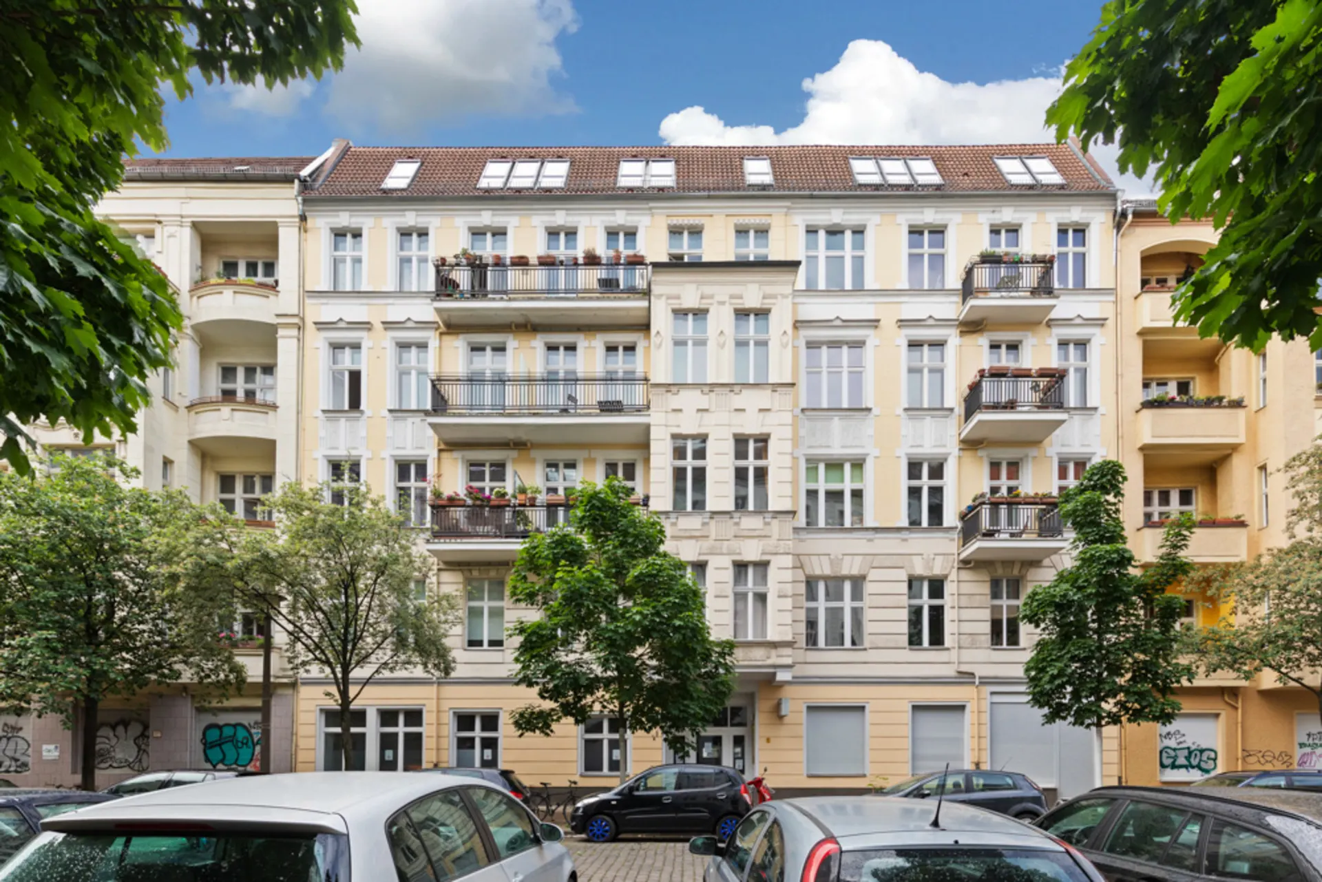 Fünfstöckiges Wohnhaus mit Balkonen und geparkten Autos davor, von Bäumen umgeben.