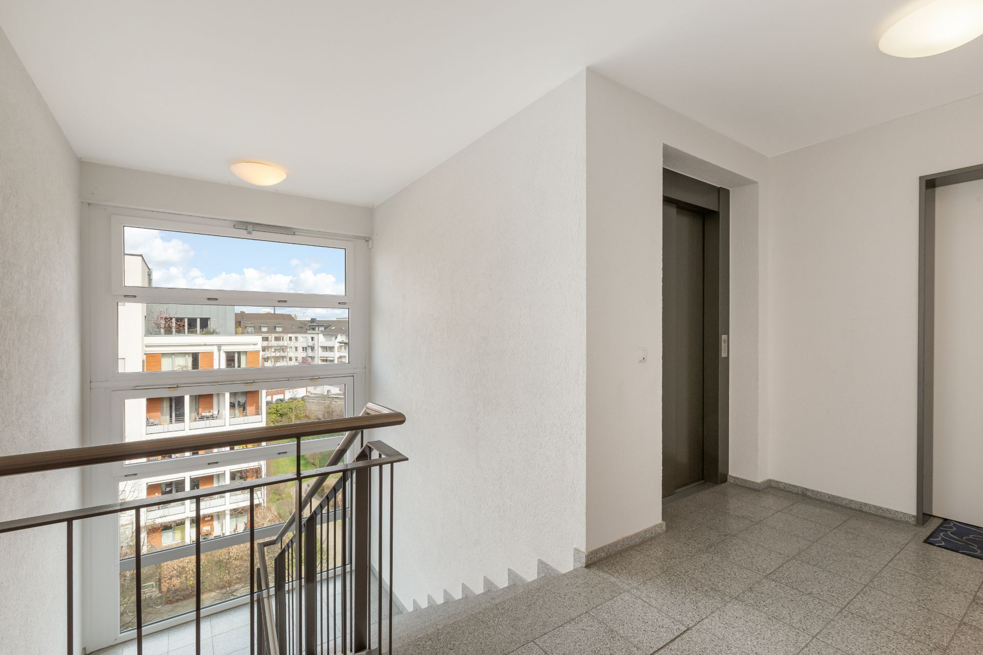 Heller Flur mit Treppengeländer, Aufzug und großem Fenster in einem Wohnhaus.
