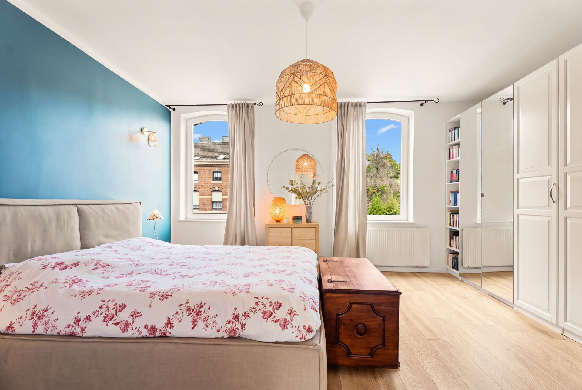 Helles Schlafzimmer mit Doppelbett, blauer Wand, Holztruhe, Kleiderschrank und zwei Fenstern.