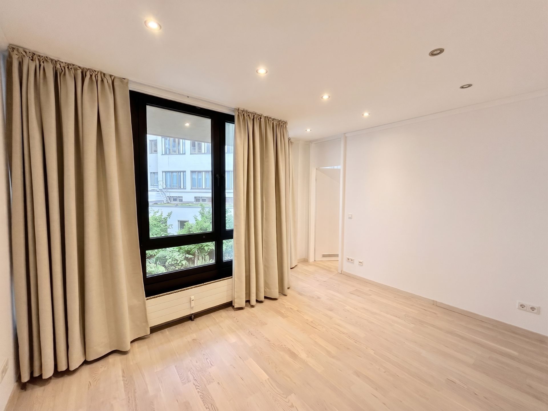 Heller, leerer Raum mit großem Fenster, beigen Vorhängen und hellem Holzboden.