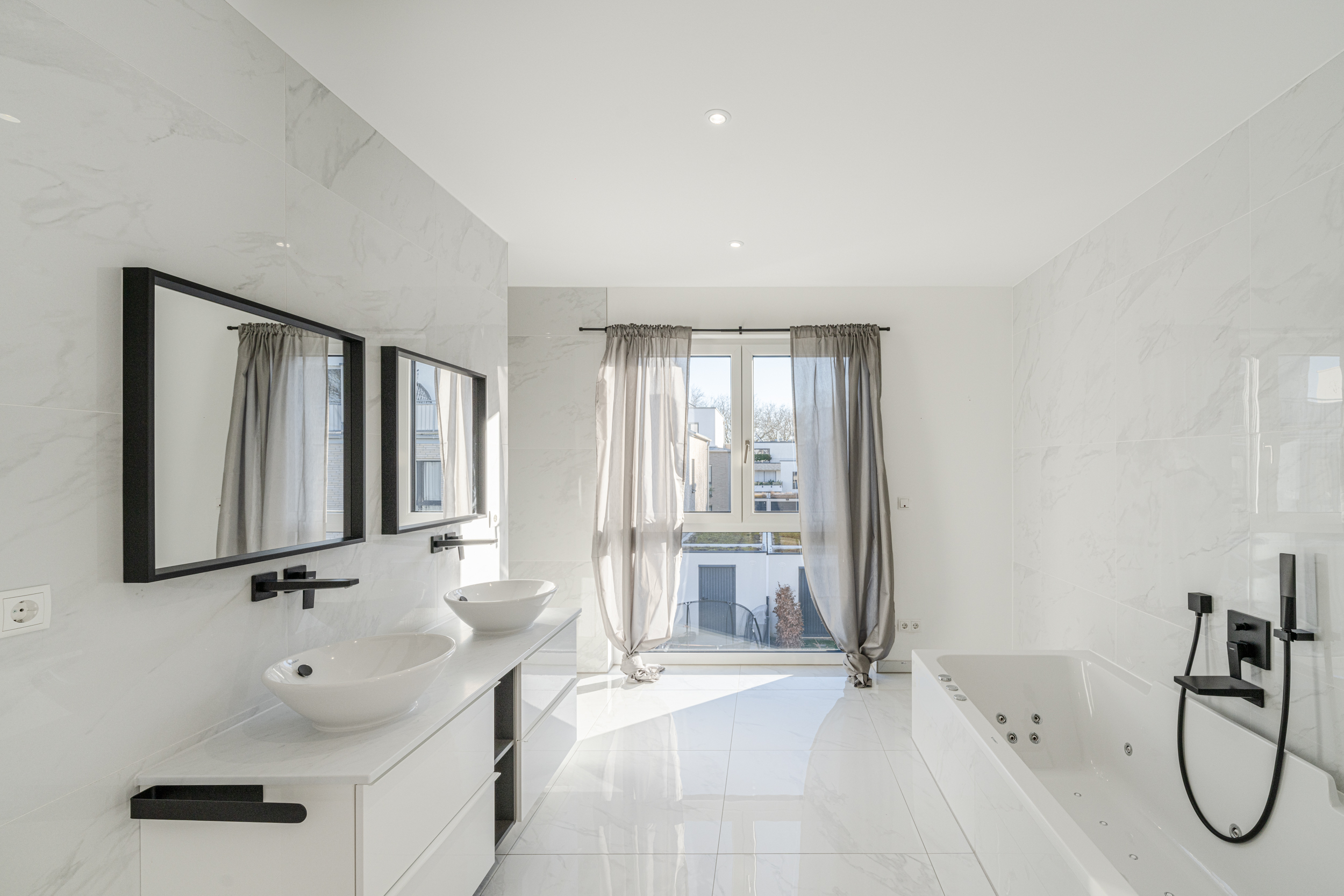 Helles Bad mit Doppelwaschbecken, großer Badewanne und Fenster mit grauen Vorhängen.