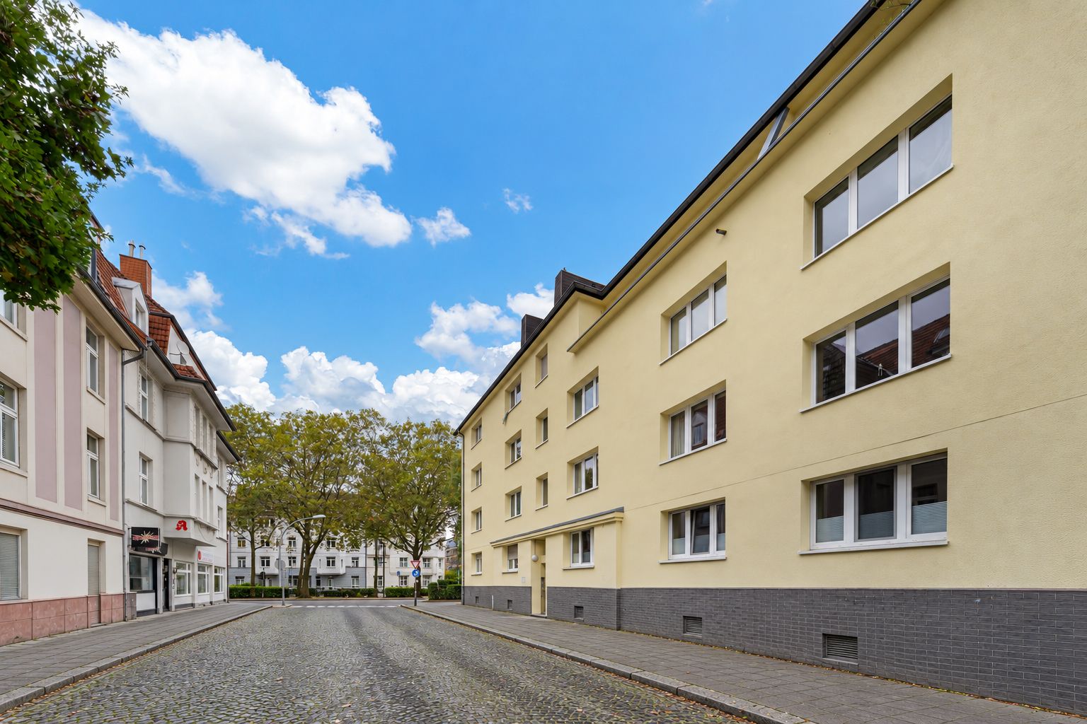 Wohnstraße mit mehrstöckigen Wohnhäusern und Kopfsteinpflaster, Bäume im Hintergrund.