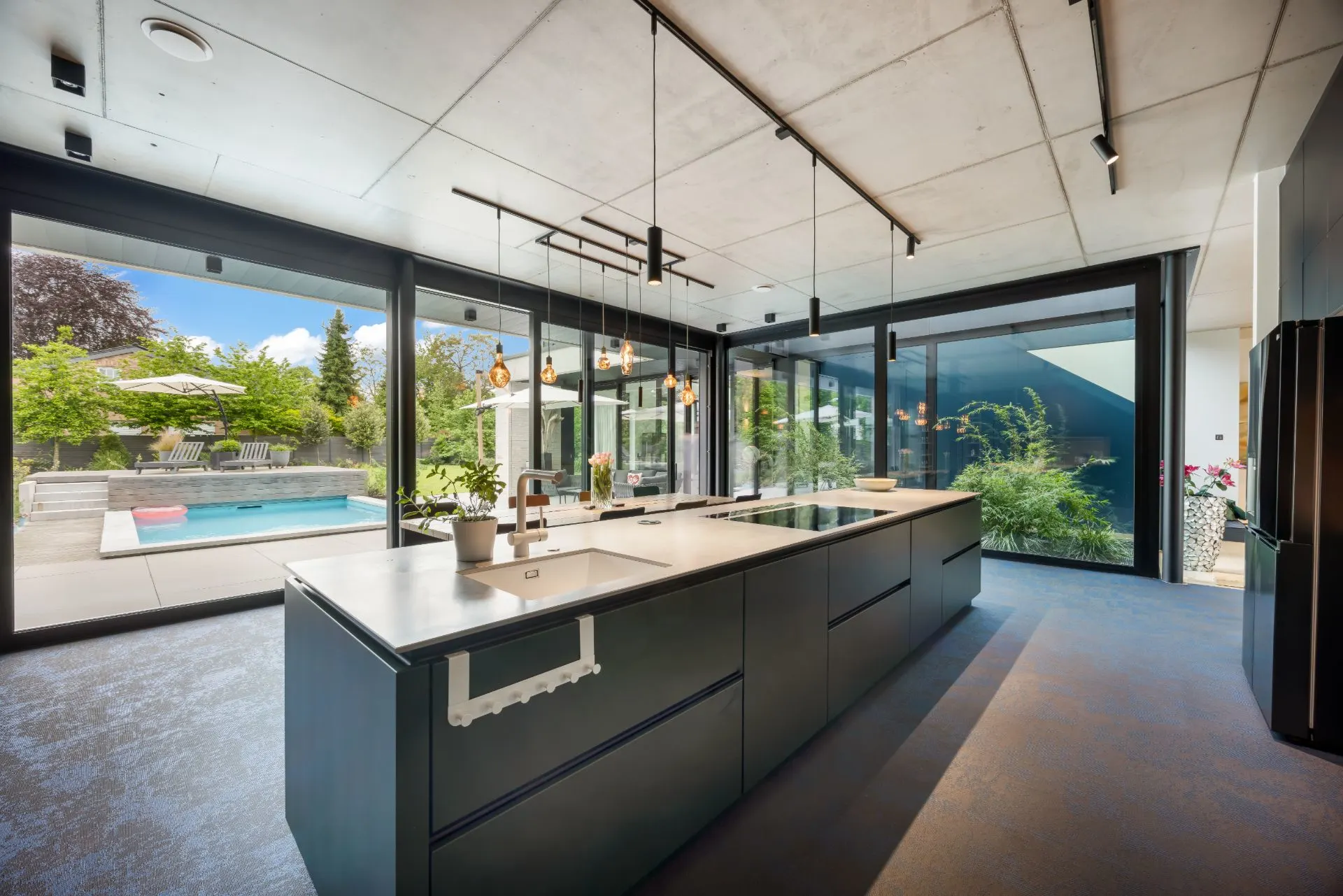 Moderne Küche mit großer Kochinsel und Blick auf Garten und Pool durch große Glasfenster.