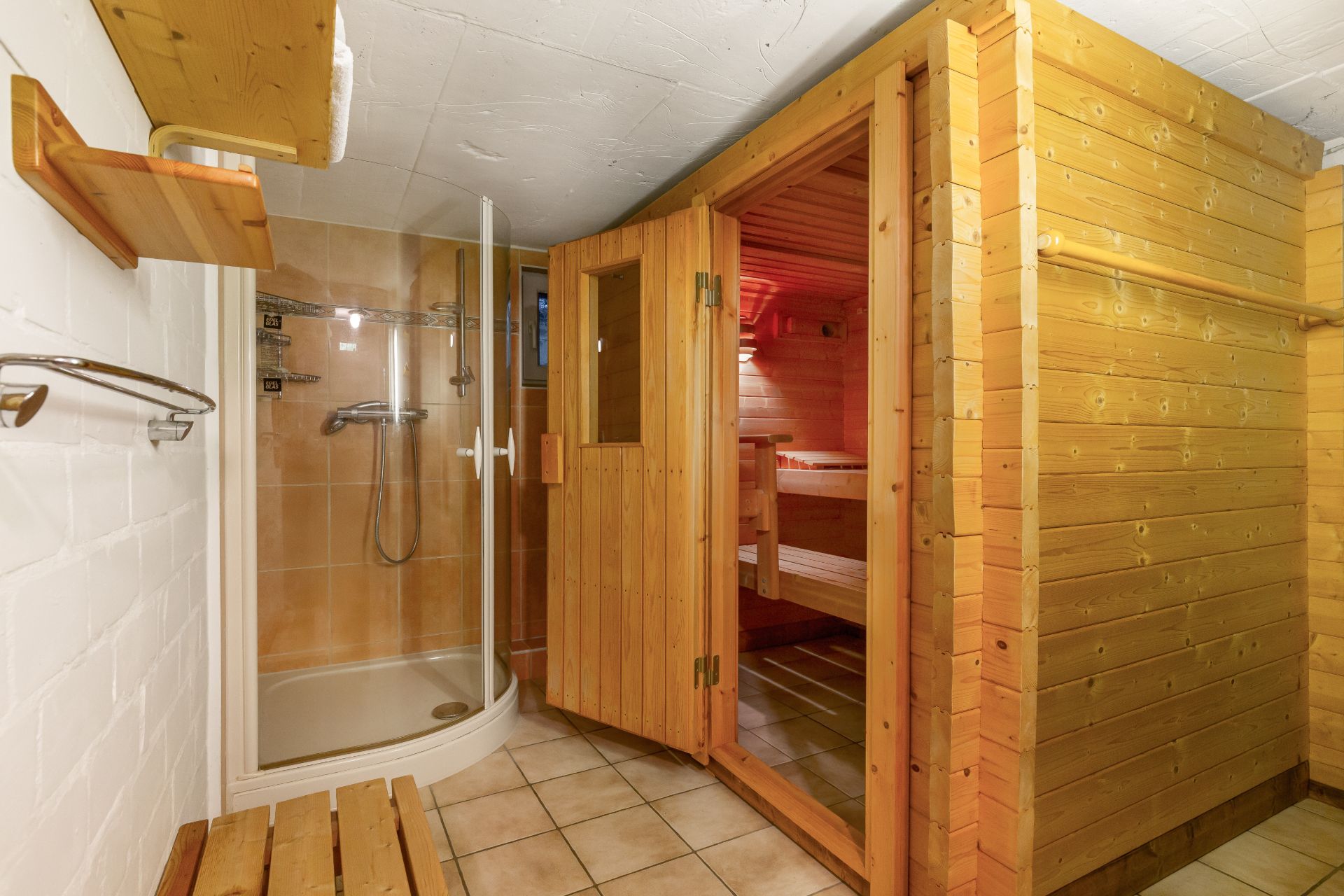 Raum mit Holzwänden, offener Sauna und Dusche, Fliesenboden und Holzbänken.