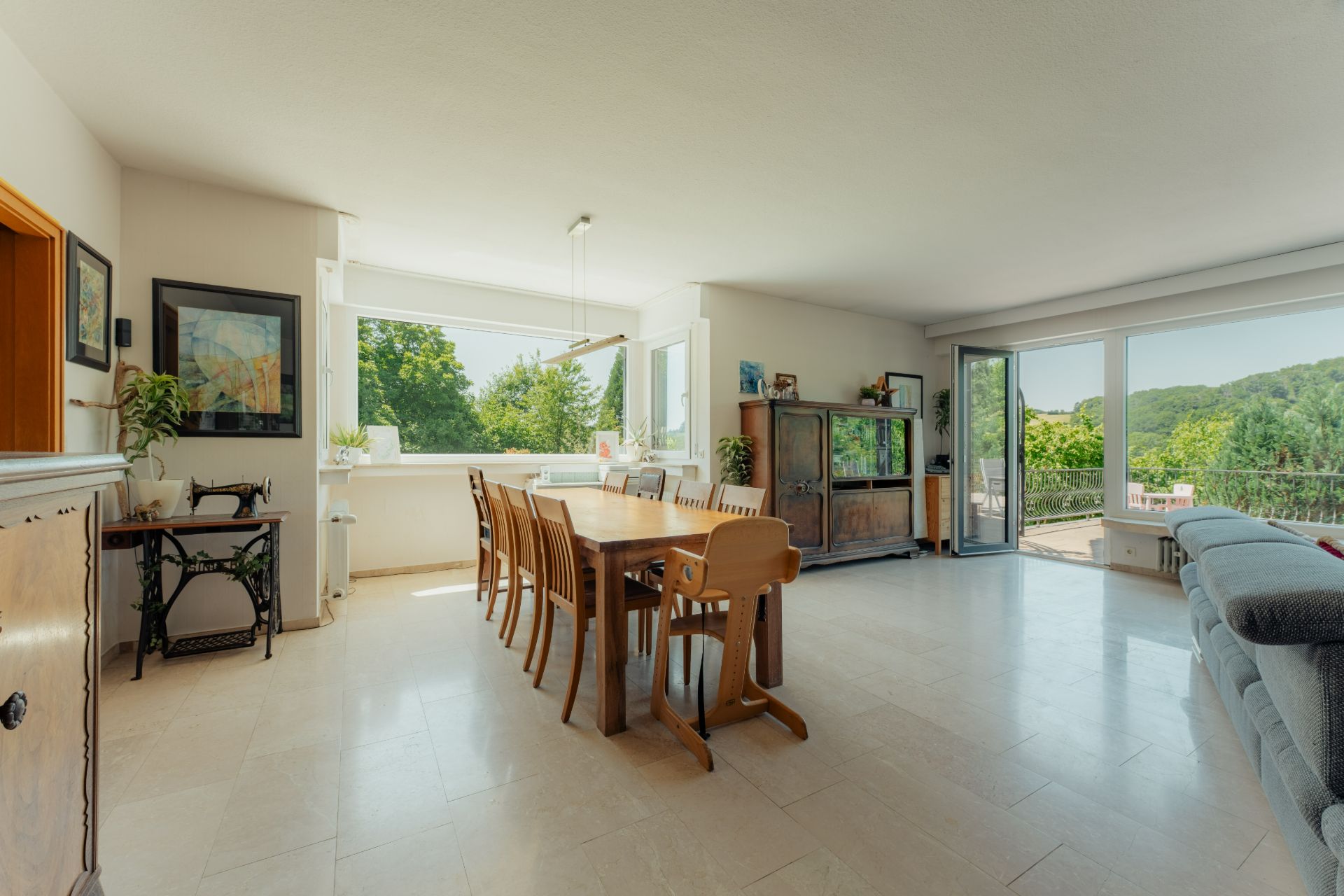 Helles Wohnzimmer mit großem Esstisch, Stühlen, Sofa und großen Fenstern mit Blick ins Grüne.