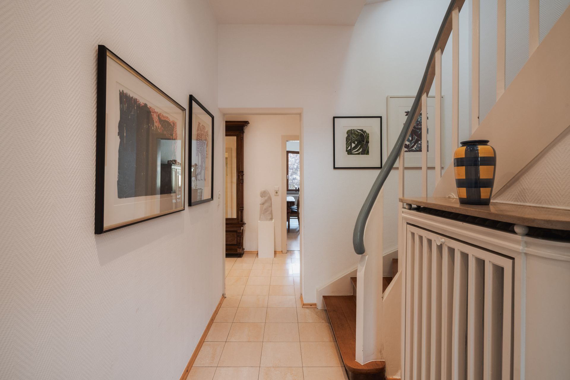 Heller Flur mit Kunst an den Wänden, Treppe und gelb-blauer Vase auf dem Geländer.
