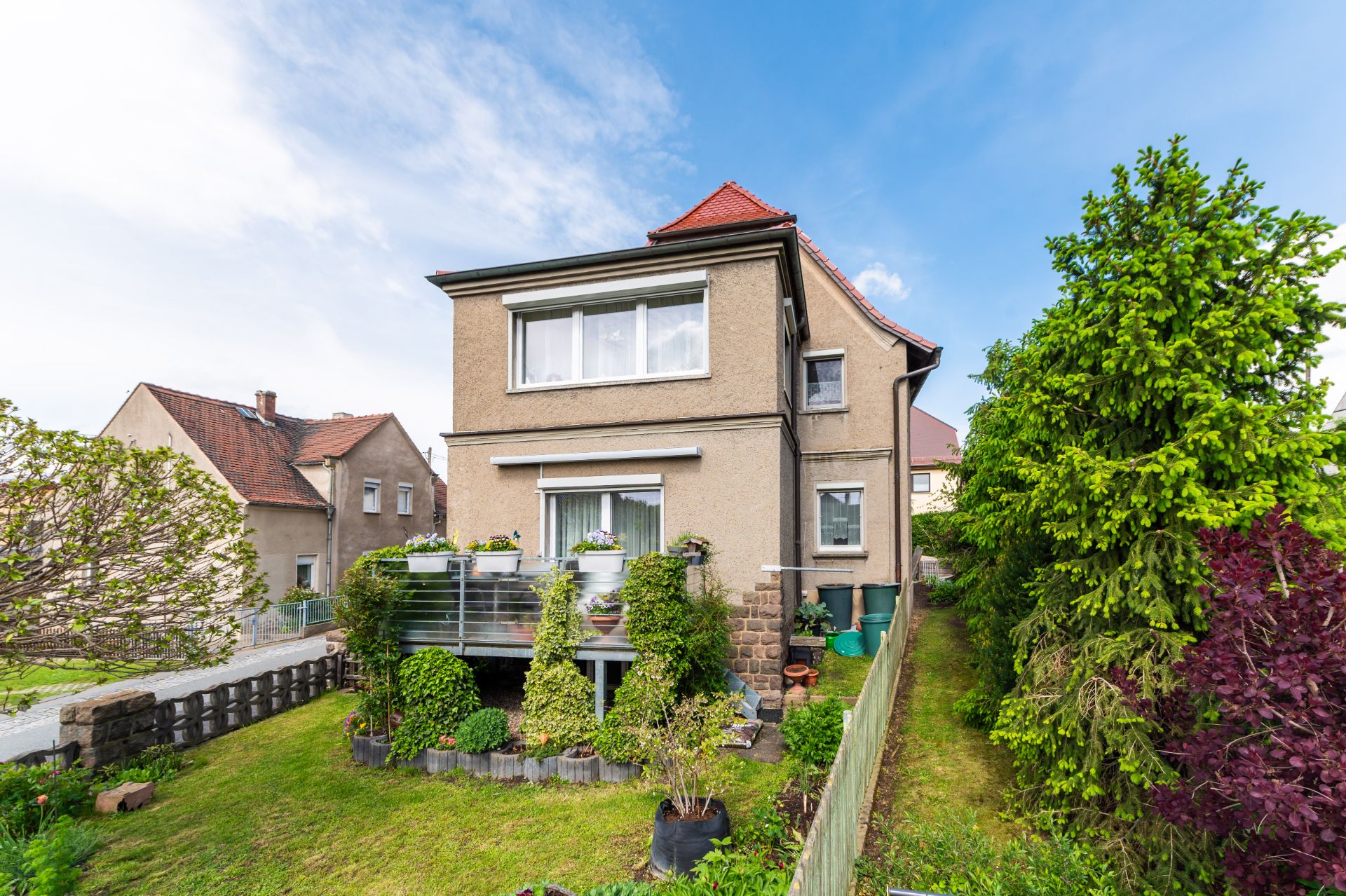 Zweistöckiges Wohnhaus mit Balkon und Garten, umgeben von Bäumen und Sträuchern.