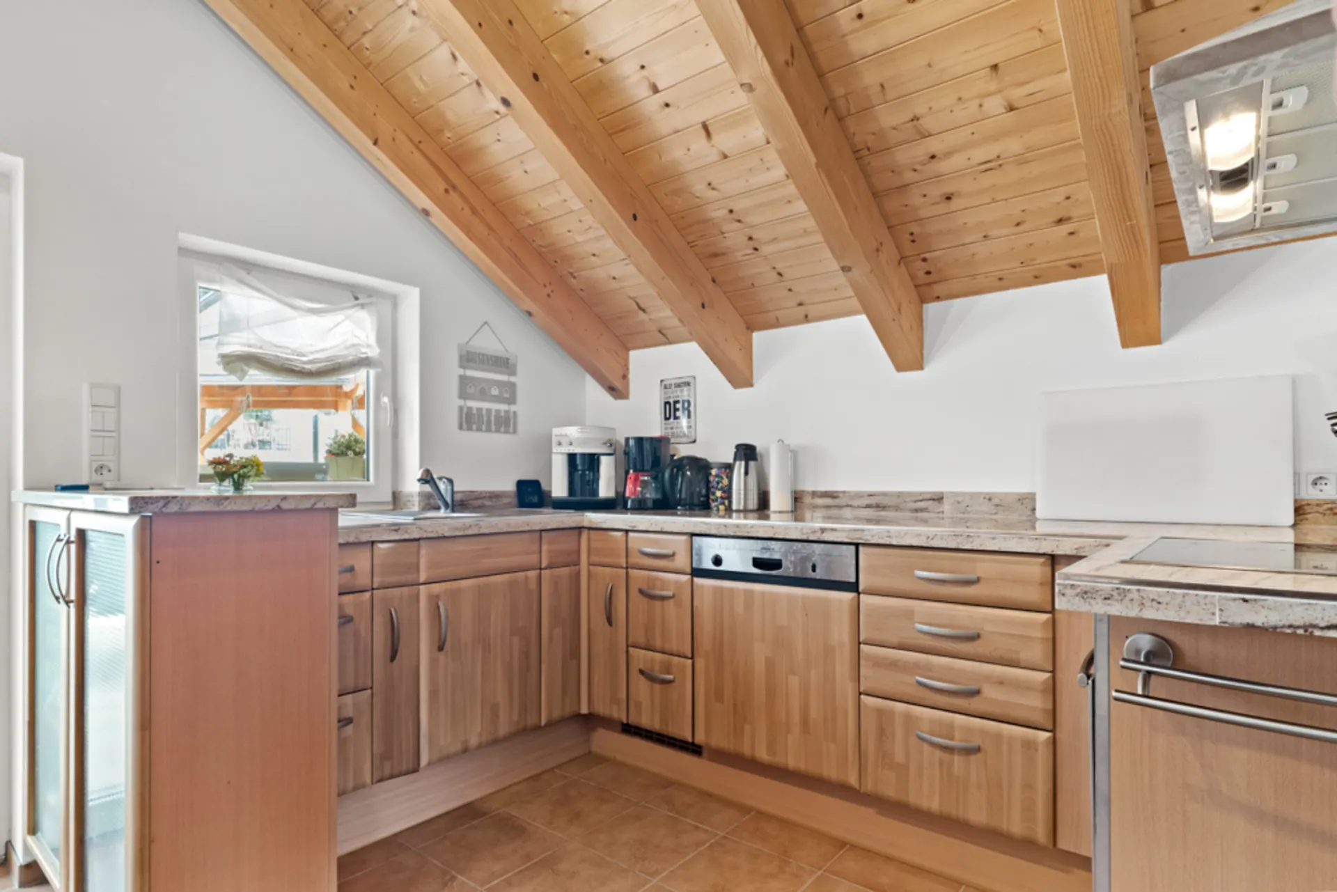 Helle Küche mit Holzfronten, Dachschräge und großem Fenster, ausgestattet mit Geräten.