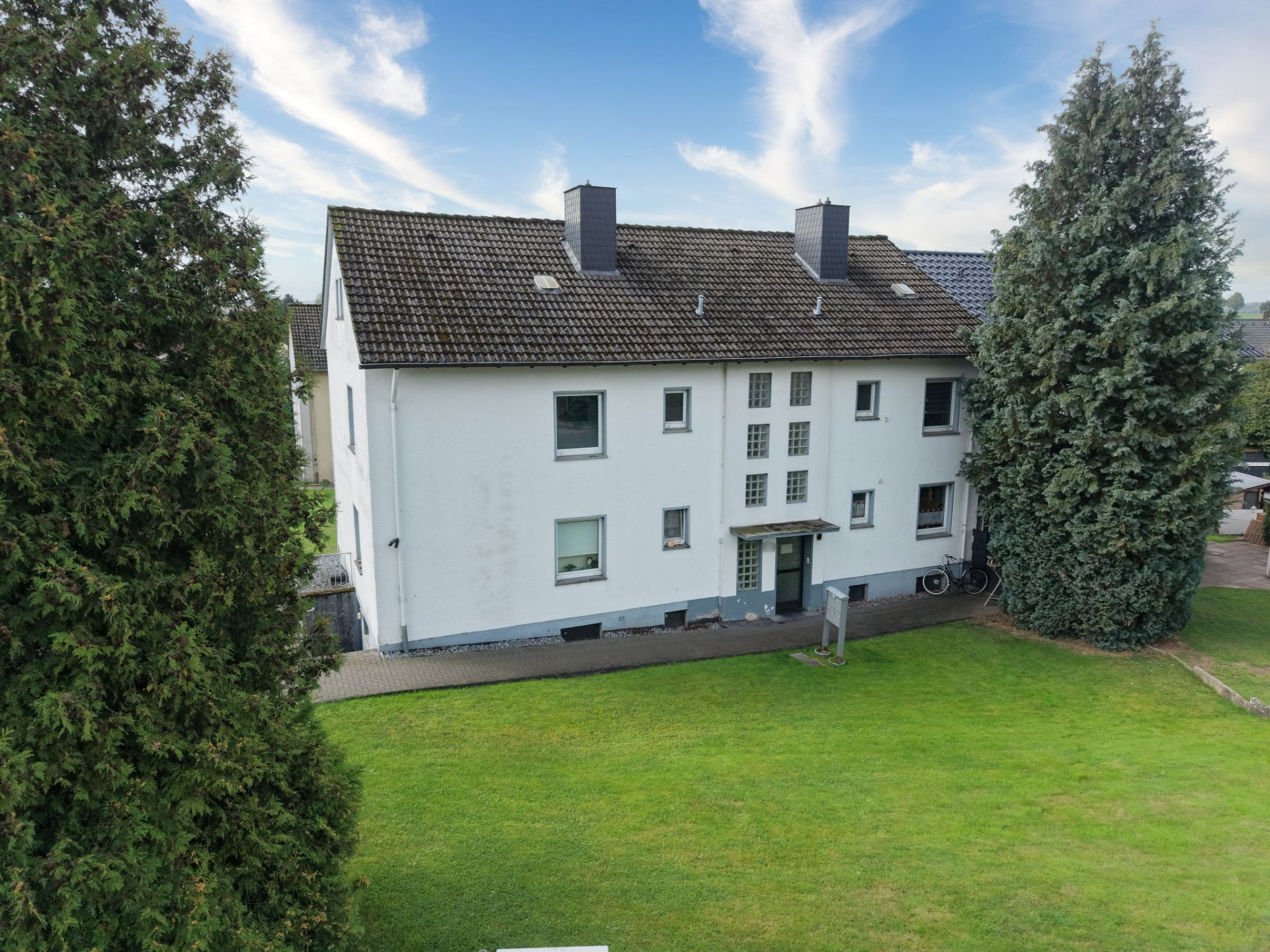 Weißes Mehrfamilienhaus mit dunklem Dach, großer Rasenfläche und Bäumen im Vordergrund.