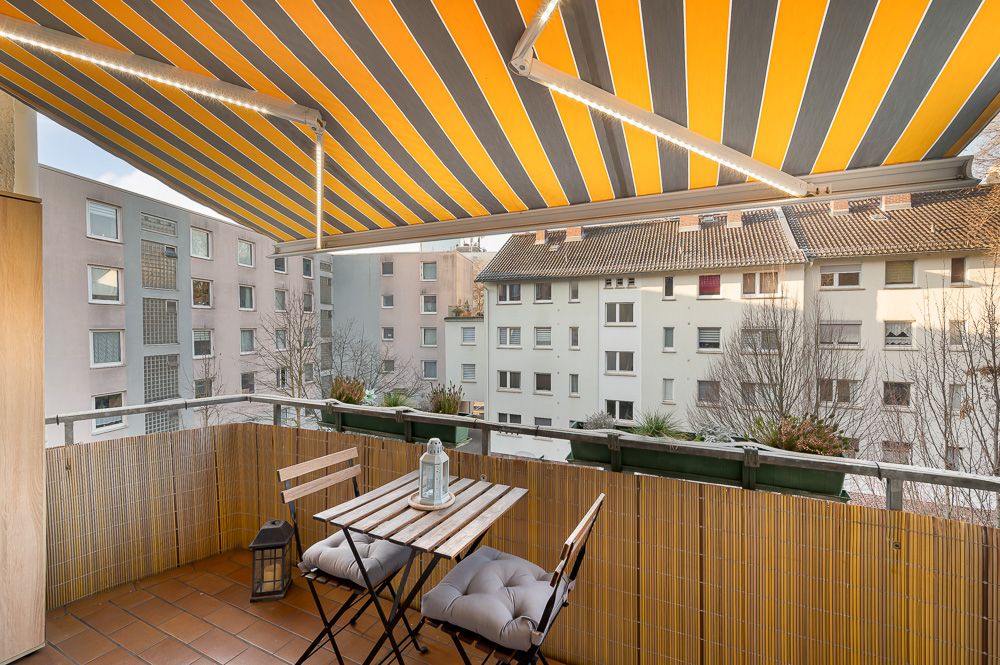 Balkon mit gelb-grauer Markise, Tisch und zwei Stühlen, Blick auf gegenüberliegende Wohnhäuser.
