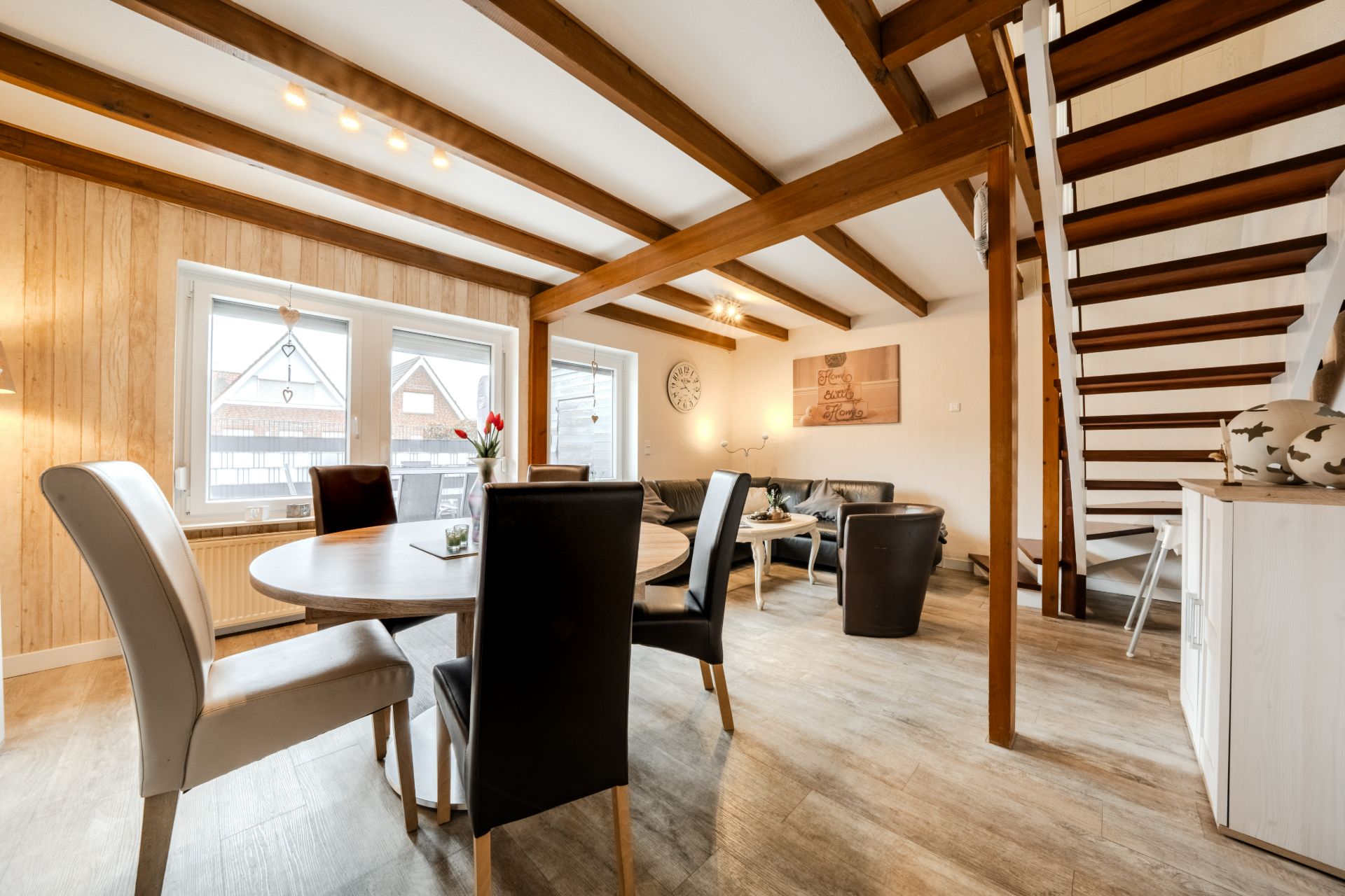Helles Wohnzimmer mit Esstisch, Sitzgruppe, Holzbalken und Treppe zu einer oberen Etage.