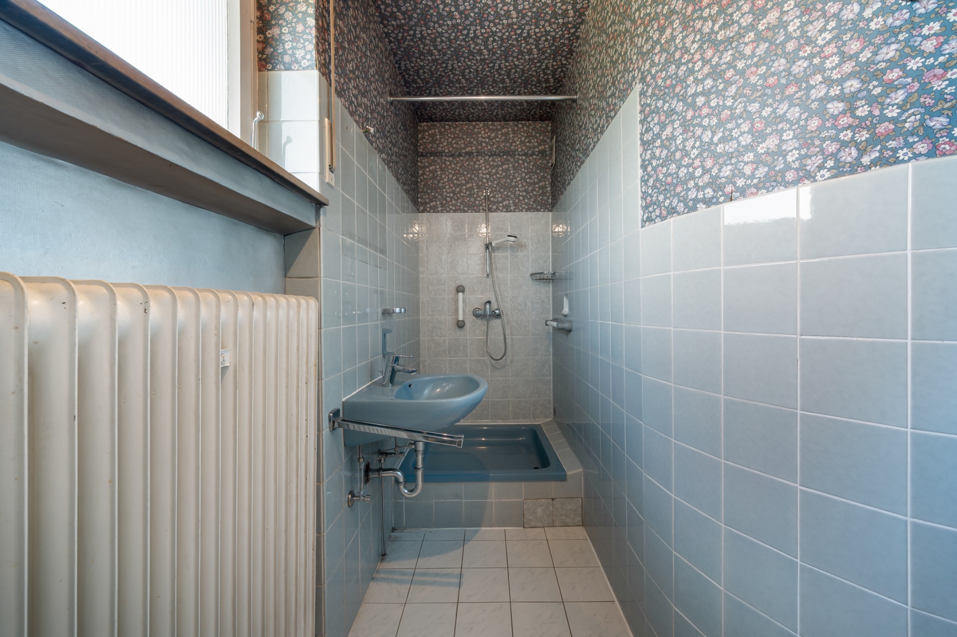 Schmales Badezimmer mit hellblauen Fliesen, Dusche, Waschbecken und Heizkörper.