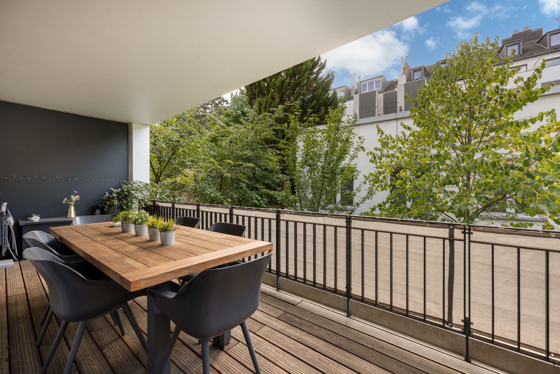 Überdachter Balkon mit Holztisch, sechs schwarzen Stühlen und Pflanzen, Blick auf Bäume und Nachbargebäude.