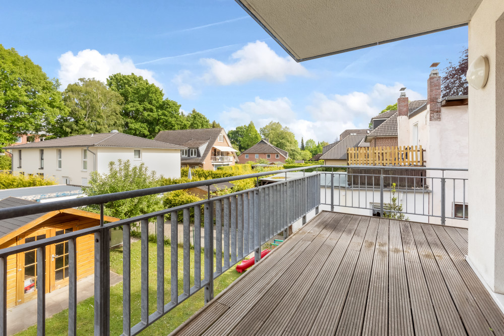 Balkon mit Holzfußboden, Ausblick auf Garten und benachbarte Wohnhäuser bei Sonnenschein.