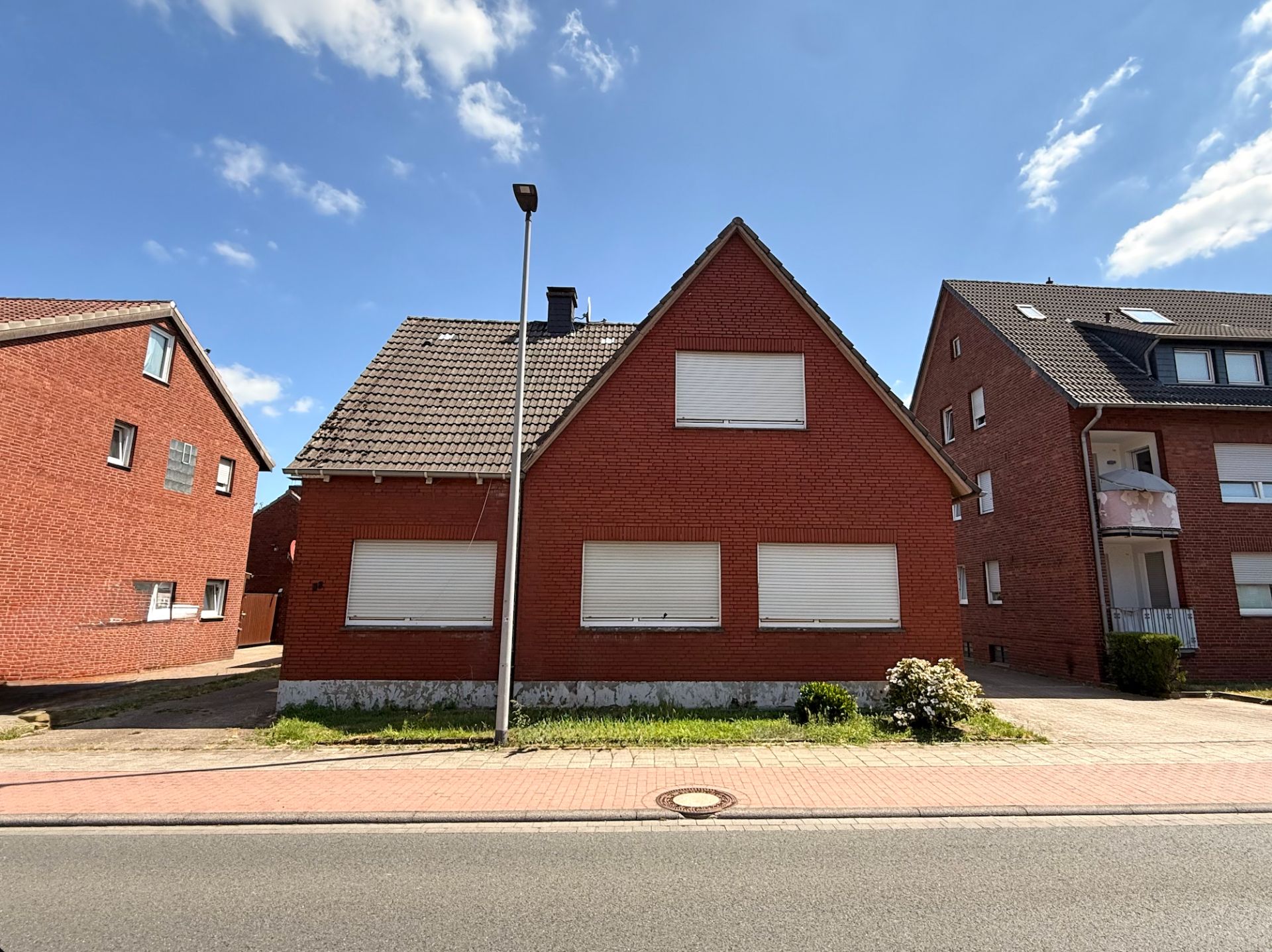 Rotes Backsteinhaus mit geschlossenen Rollläden, Vorgarten und Gehweg bei sonnigem Wetter.