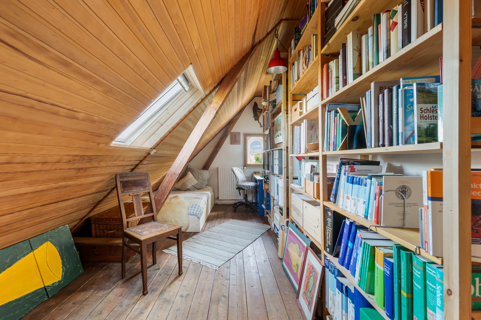 Dachzimmer mit schräger Holzdecke, Bücherregalen, Stuhl und kleinem Sofa am Fenster.