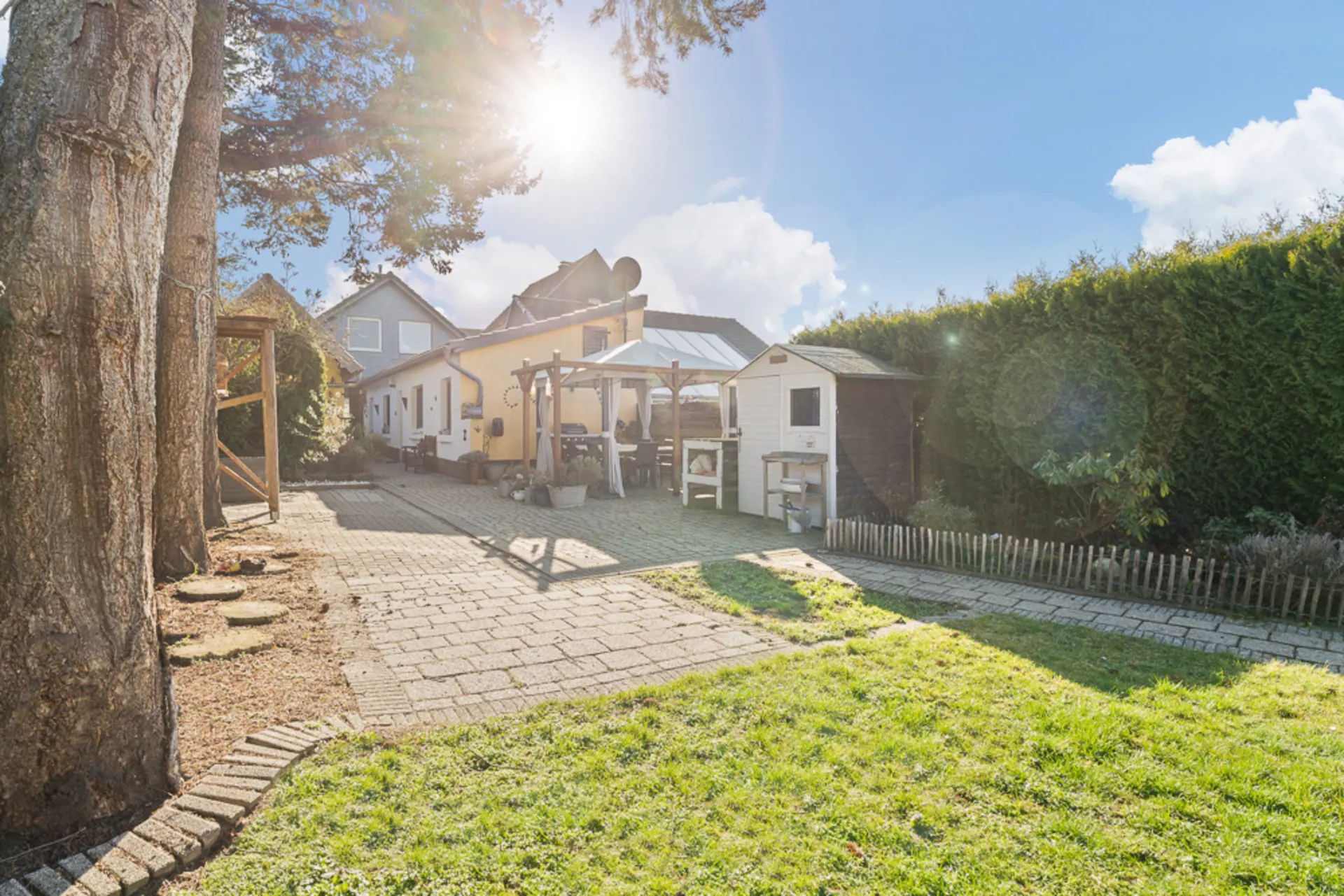 Garten mit Rasen, Terrasse, Pavillon, Baum, Gartenhaus und Hecke bei Sonnenschein.