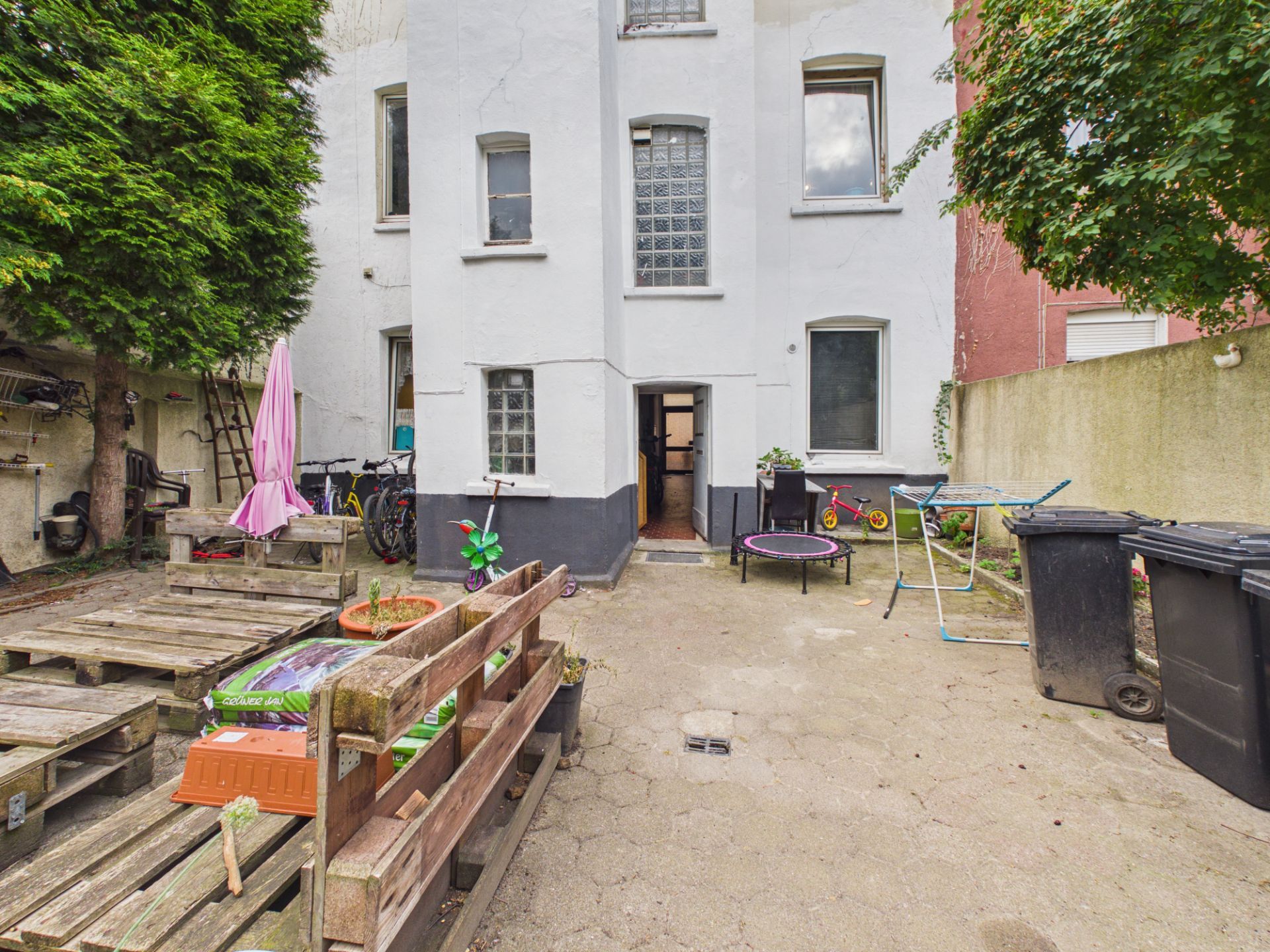 Hinterhof eines Wohnhauses mit Palettenbank, Fahrrädern, Spielzeug und Mülltonnen.