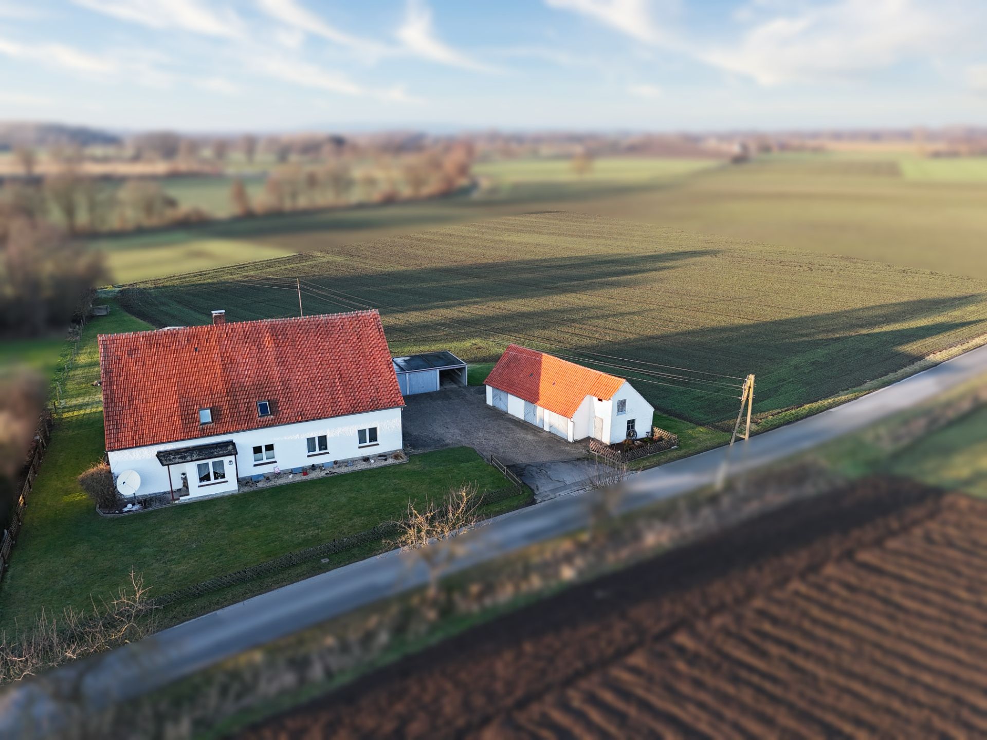 Luftaufnahme eines weißen Hauses mit rotem Dach und Nebengebäude auf einem großen Grundstück am Feldrand.