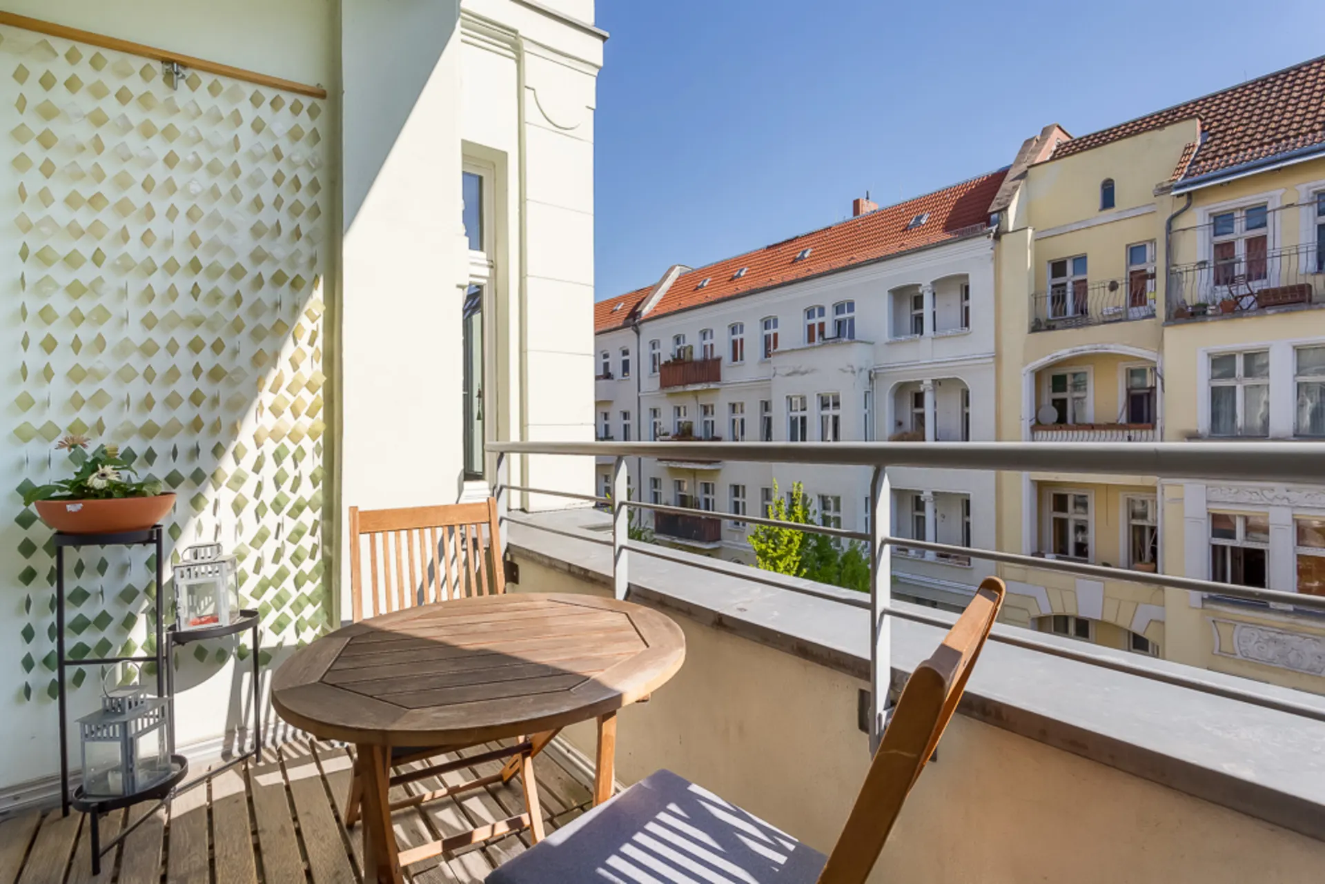 Balkon mit Holztisch und zwei Stühlen, Pflanze auf Regal, Blick auf Wohnhäuser.