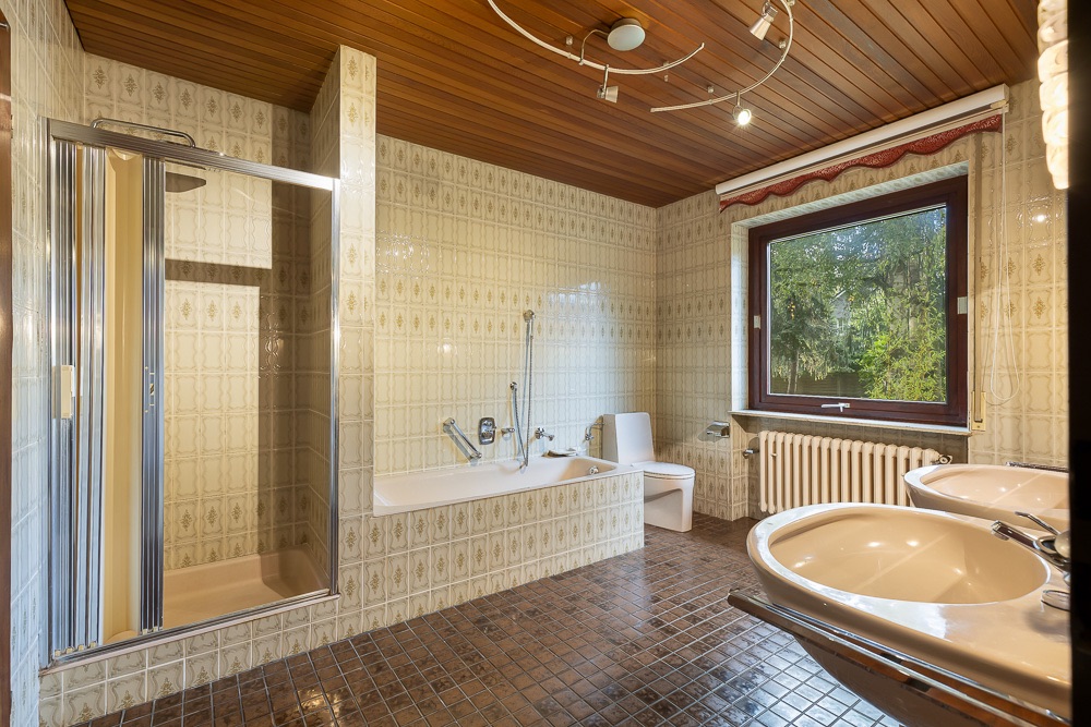 Großes Badezimmer mit Dusche, Badewanne, Toilette, zwei Waschbecken und Fenster mit Blick ins Grüne.