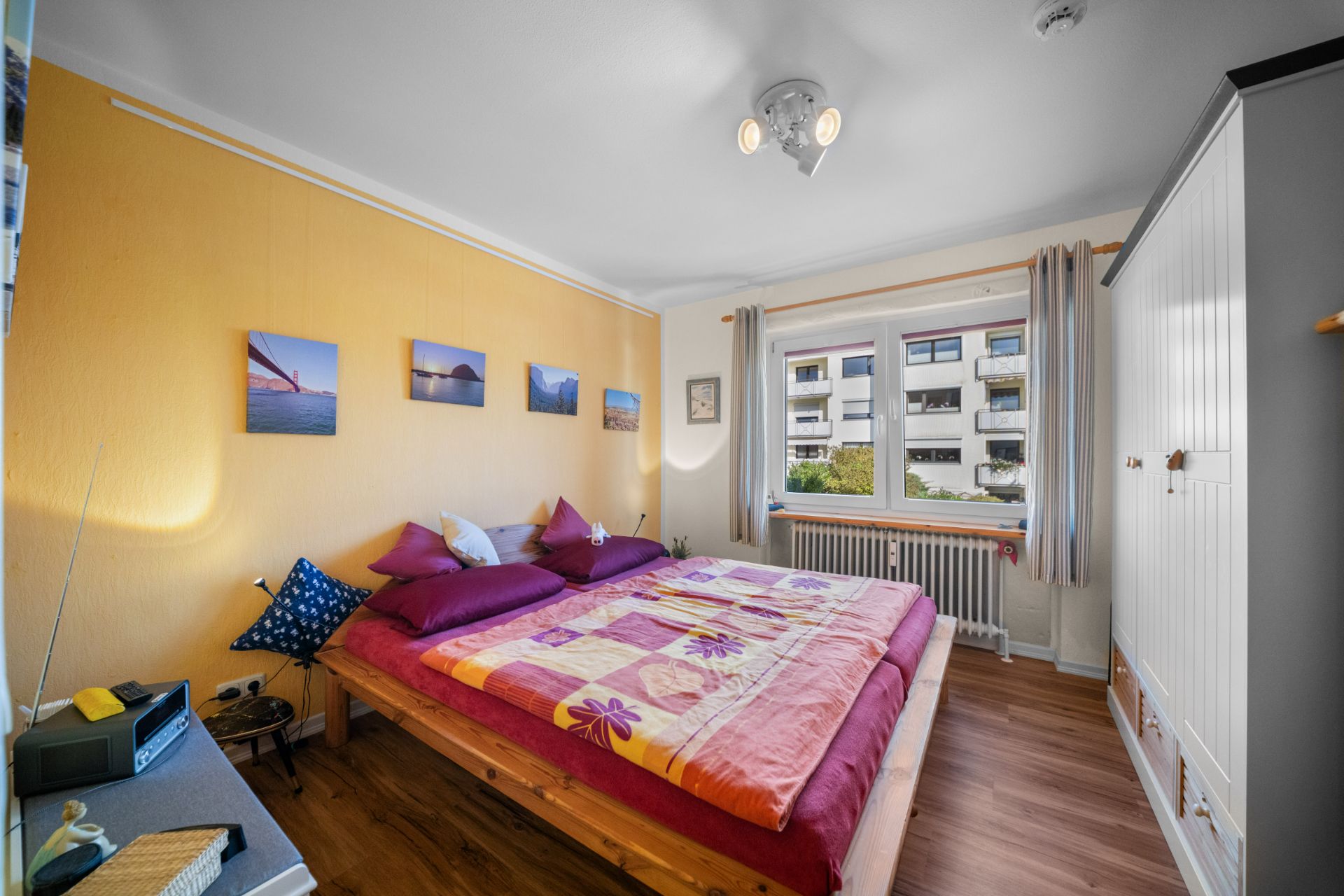 Helles Schlafzimmer mit Doppelbett, buntem Bettbezug, Kleiderschrank und großem Fenster.