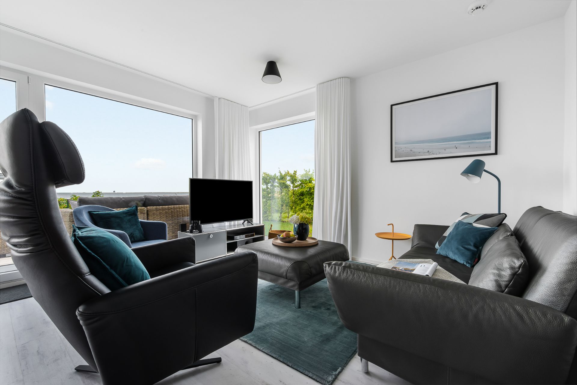 Modernes Wohnzimmer mit schwarzem Ledersofa, Sessel, TV und großen Fenstern mit Ausblick.