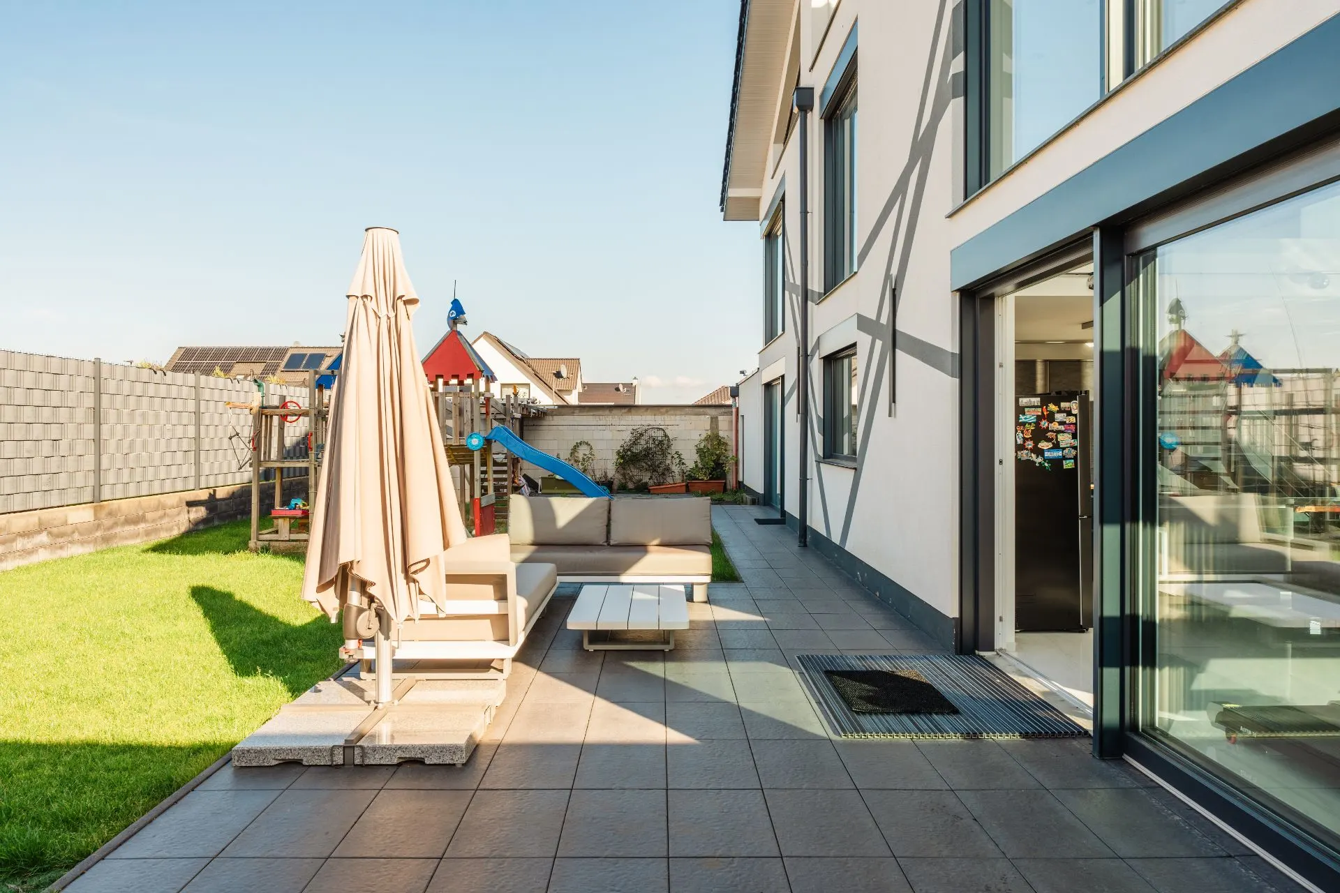 Moderne Terrasse mit Sofa, Sonnenschirm und Spielplatz im Garten neben einem Wohnhaus.