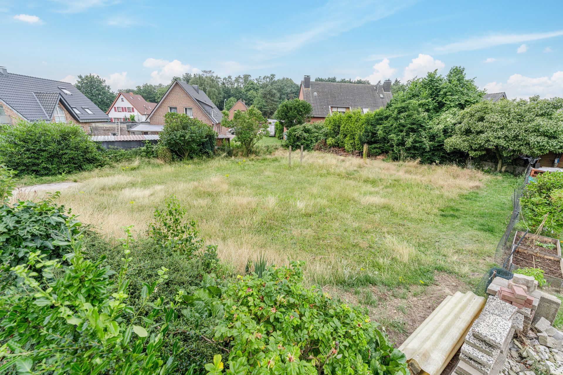 Leeres, grasbewachsenes Grundstück mit Sträuchern und angrenzenden Wohnhäusern.