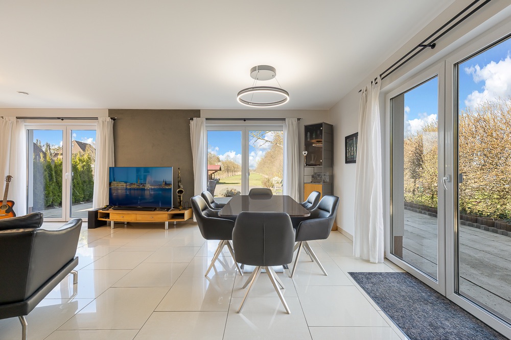 Helles Wohnzimmer mit Esstisch, Fernseher, Sesseln und großen Fenstern zum Garten.