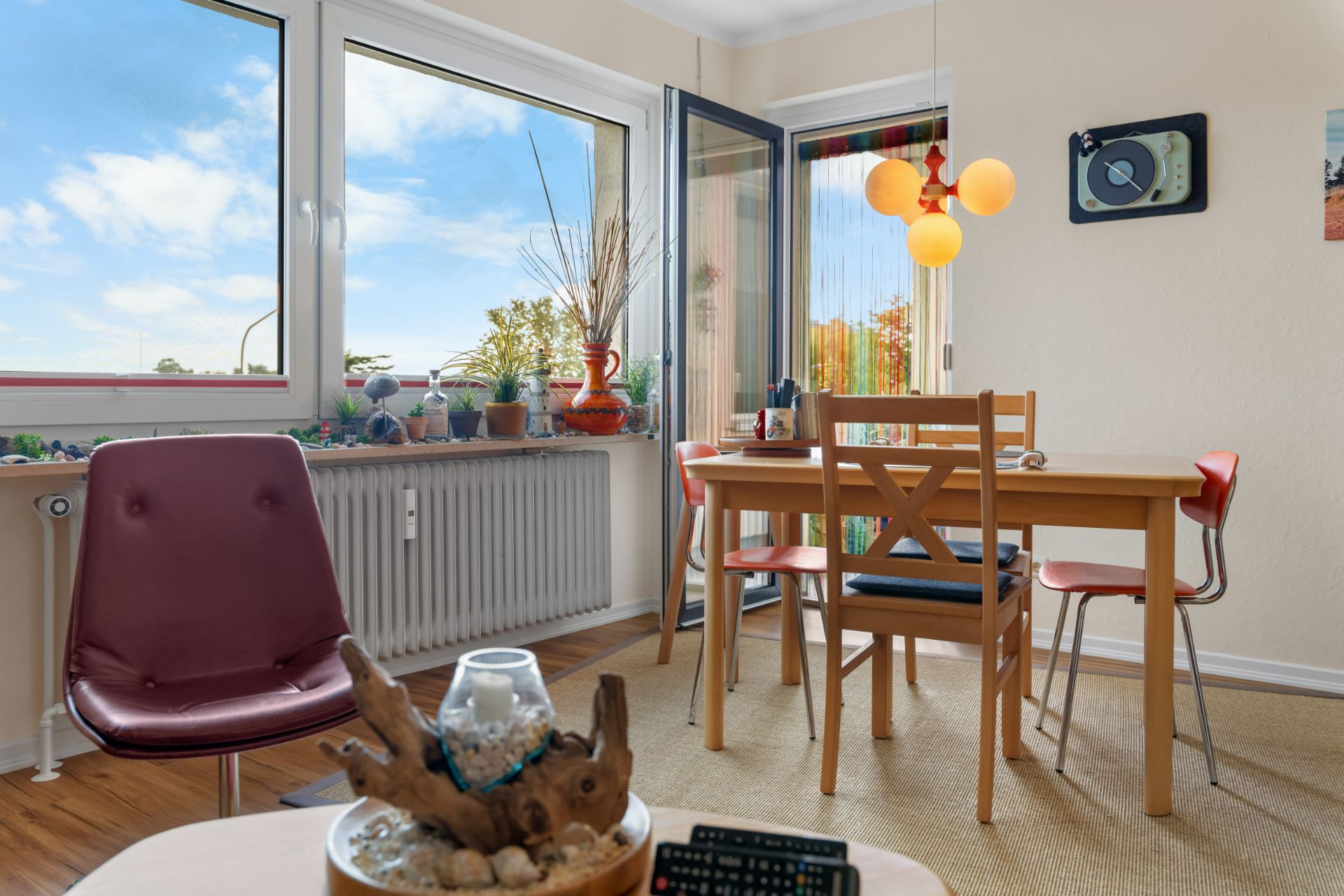 Essbereich mit Holztisch, vier Stühlen, großen Fenstern und offener Balkontür, Pflanzen auf der Fensterbank.