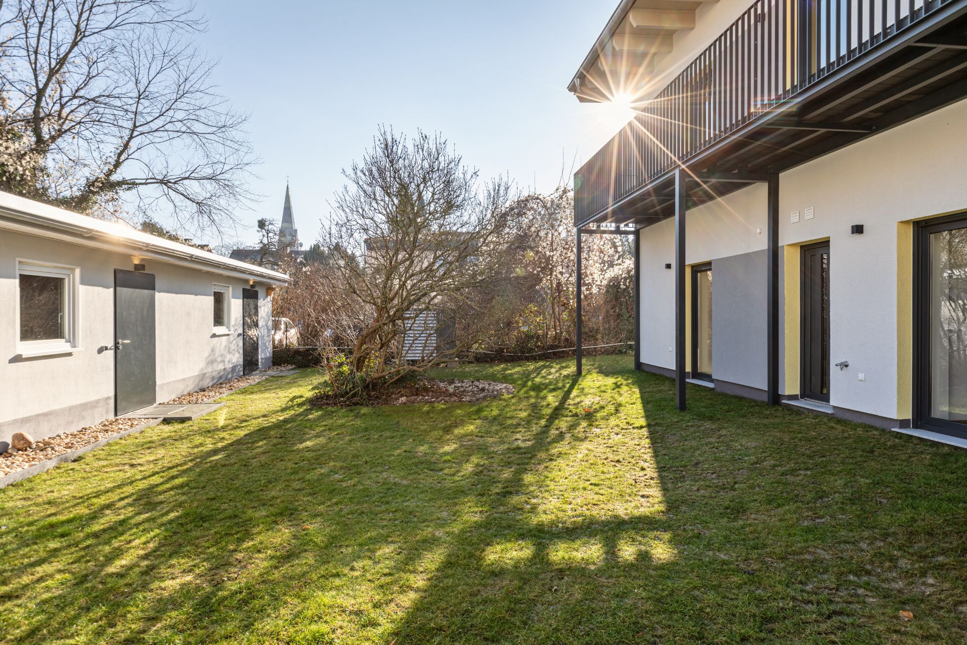 Garten mit Rasenfläche, Strauch, modernem Haus und Nebengebäude bei Sonnenschein.