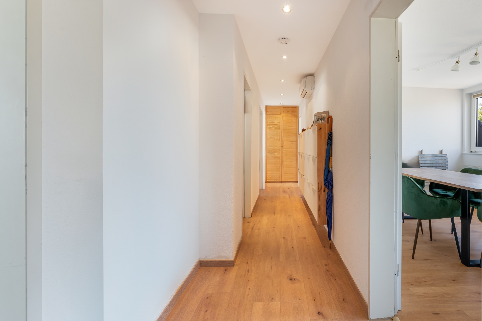 Langer Flur mit Holzfußboden und weißen Wänden, rechts Zugang zu einem Esszimmer mit grünen Stühlen.