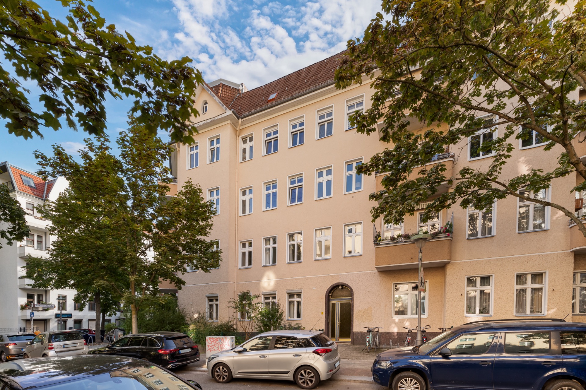 Mehrstöckiges Wohngebäude mit beigen Fassaden, Bäumen davor und geparkten Autos am Straßenrand.