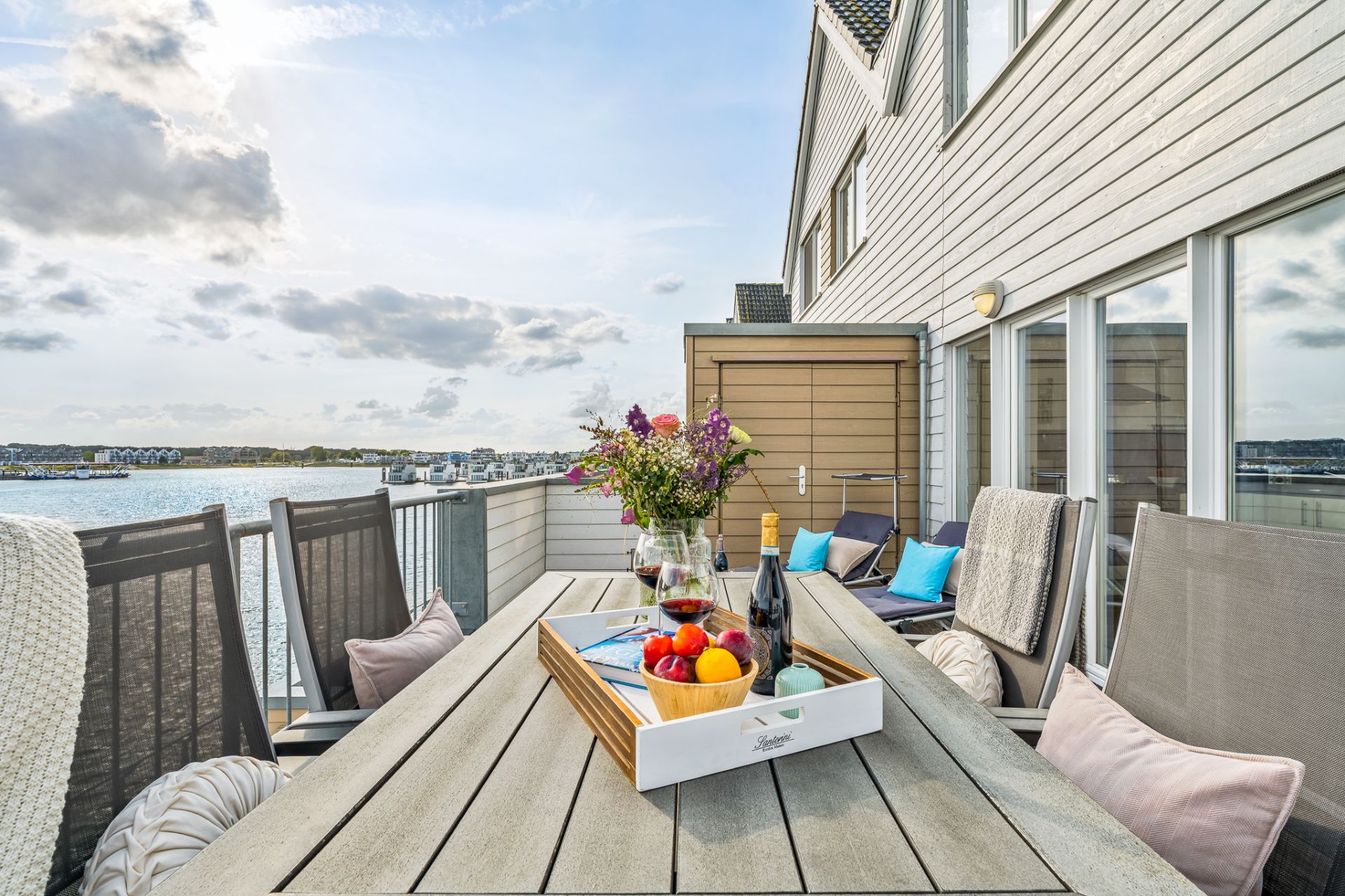 Balkon mit Esstisch, Stühlen, Blumen und Getränken, Ausblick auf Wasser und Häuser.