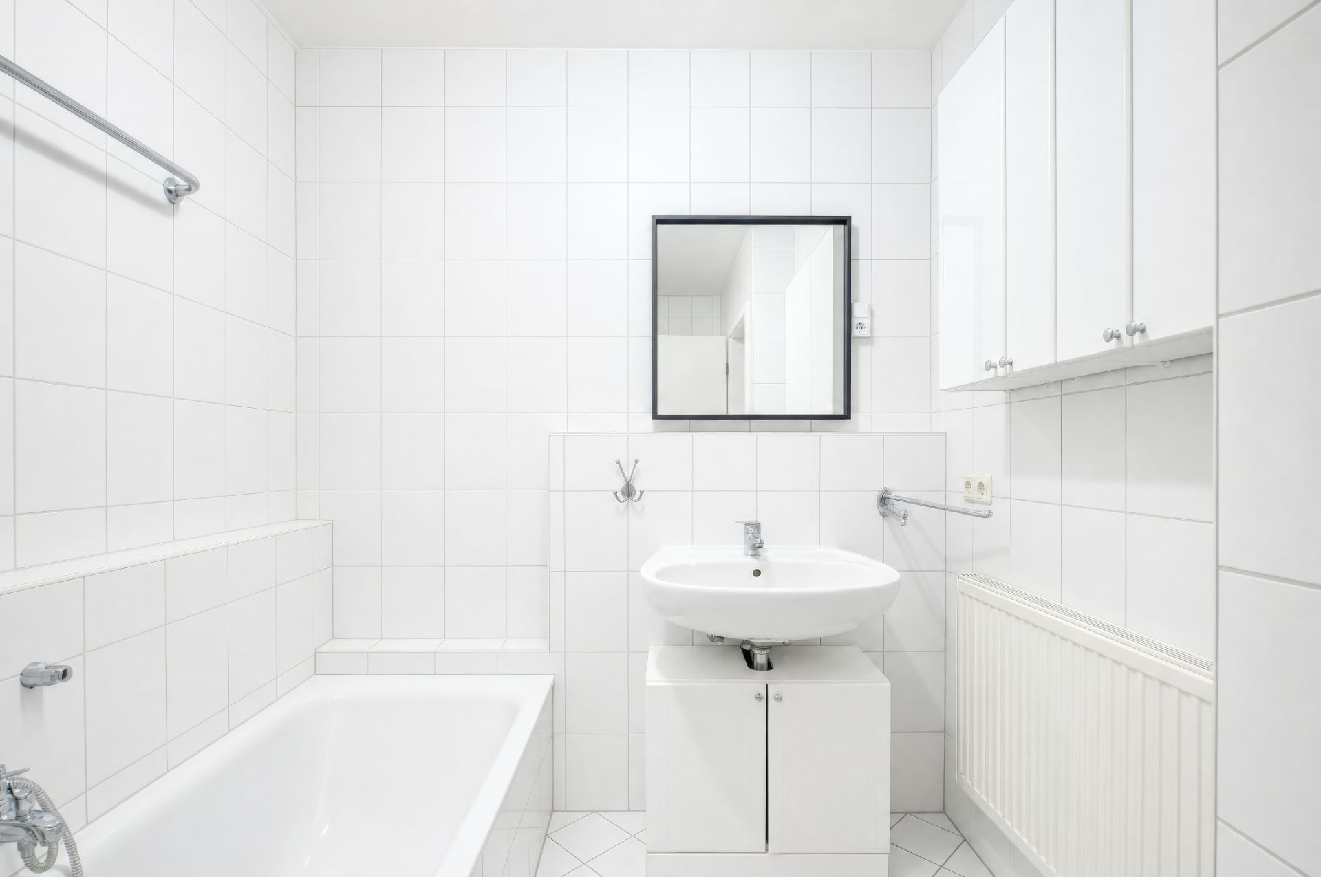 Weiß gefliestes Badezimmer mit Badewanne, Waschbecken, Spiegel und Hängeschränken.