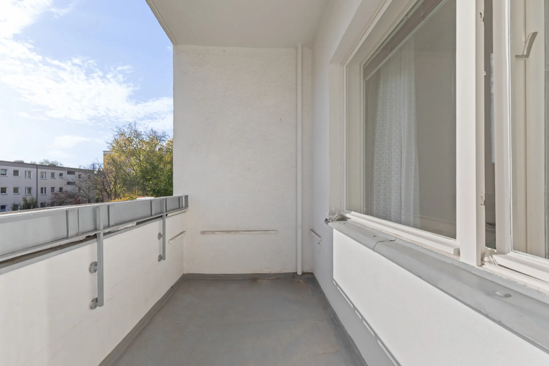 Ein leerer Balkon mit weißem Geländer und angrenzendem Fenster, Blick auf Bäume und Nachbargebäude.
