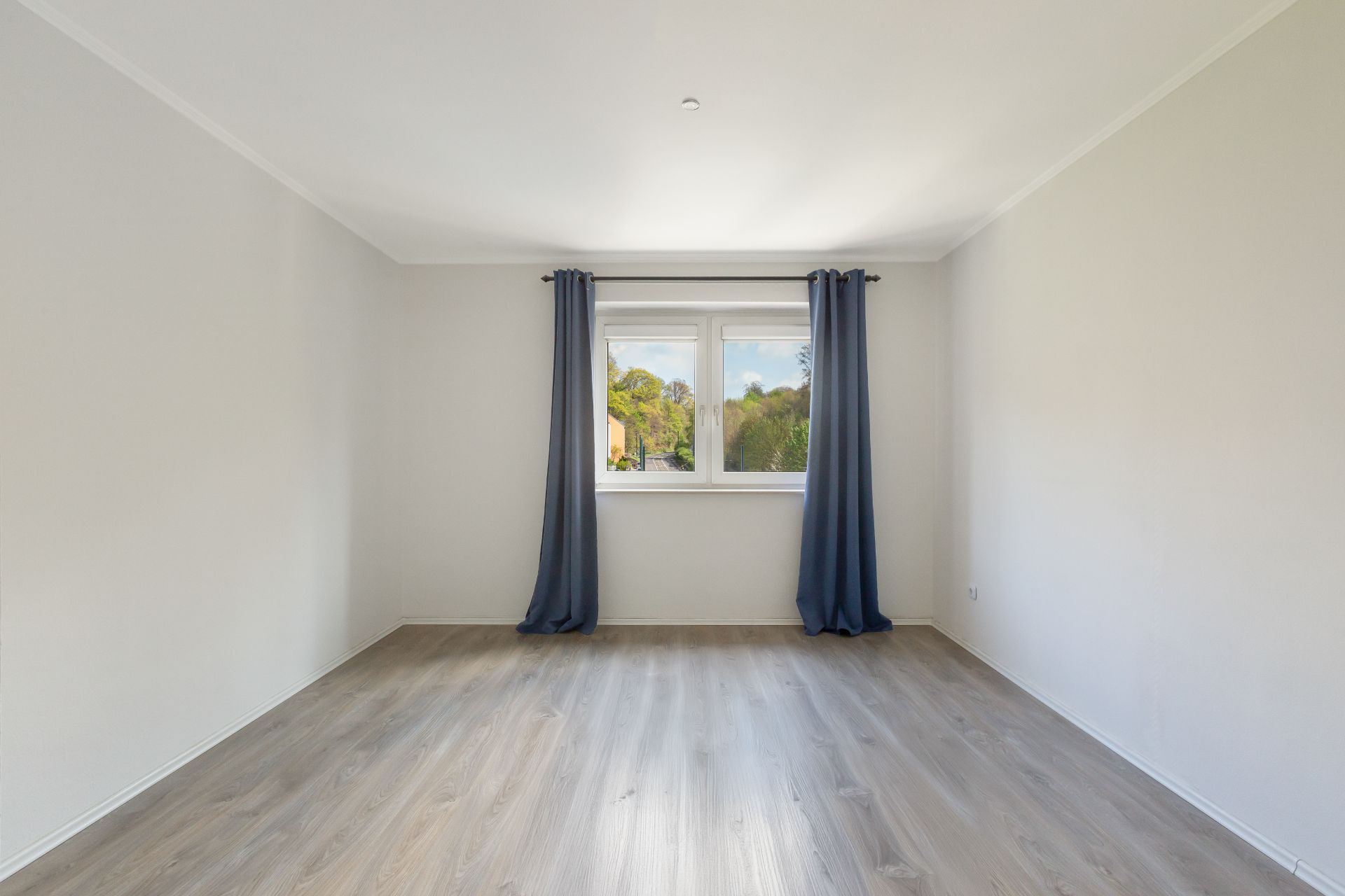 Leeres Zimmer mit hellen Wänden, Holzfußboden und Fenster mit blauen Vorhängen.