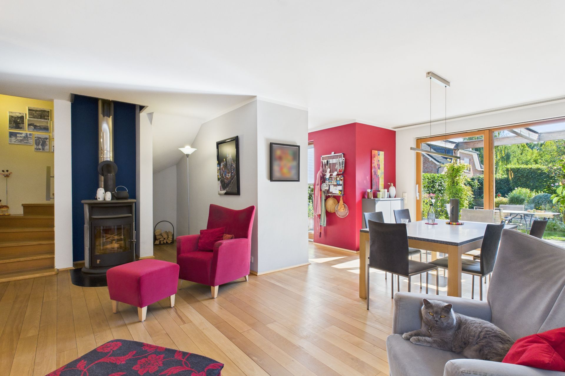 Helles Wohnzimmer mit Holzfußboden, Kamin, roten Sesseln, Esstisch und großer Fensterfront zum Garten.