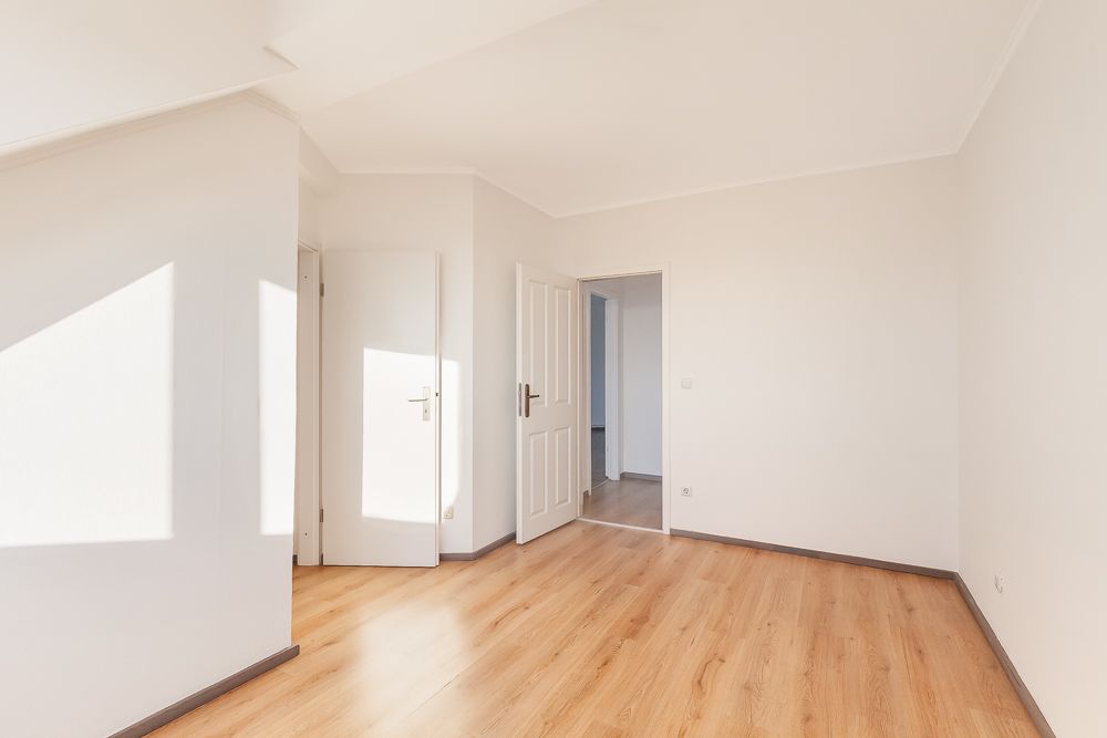 Helles, leeres Zimmer mit Holzboden und weißen Wänden, zwei offene Türen.