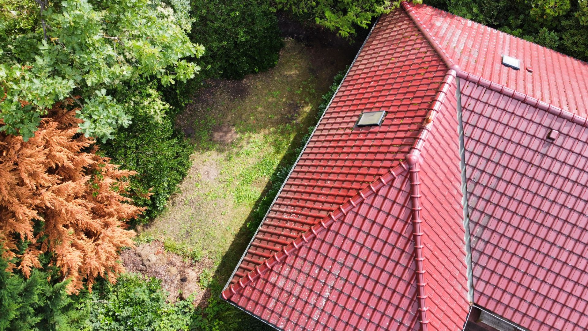 Dach eines Hauses mit roten Ziegeln, umgeben von Bäumen und etwas Rasenfläche.