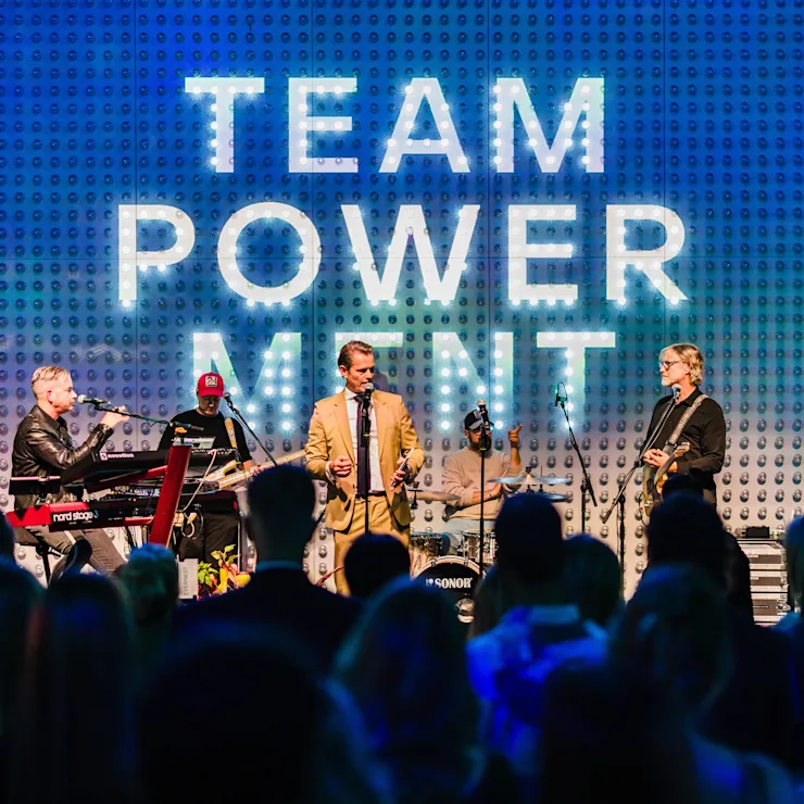 Eine Band performt auf einer Bühne vor großem Schriftzug und Publikum.