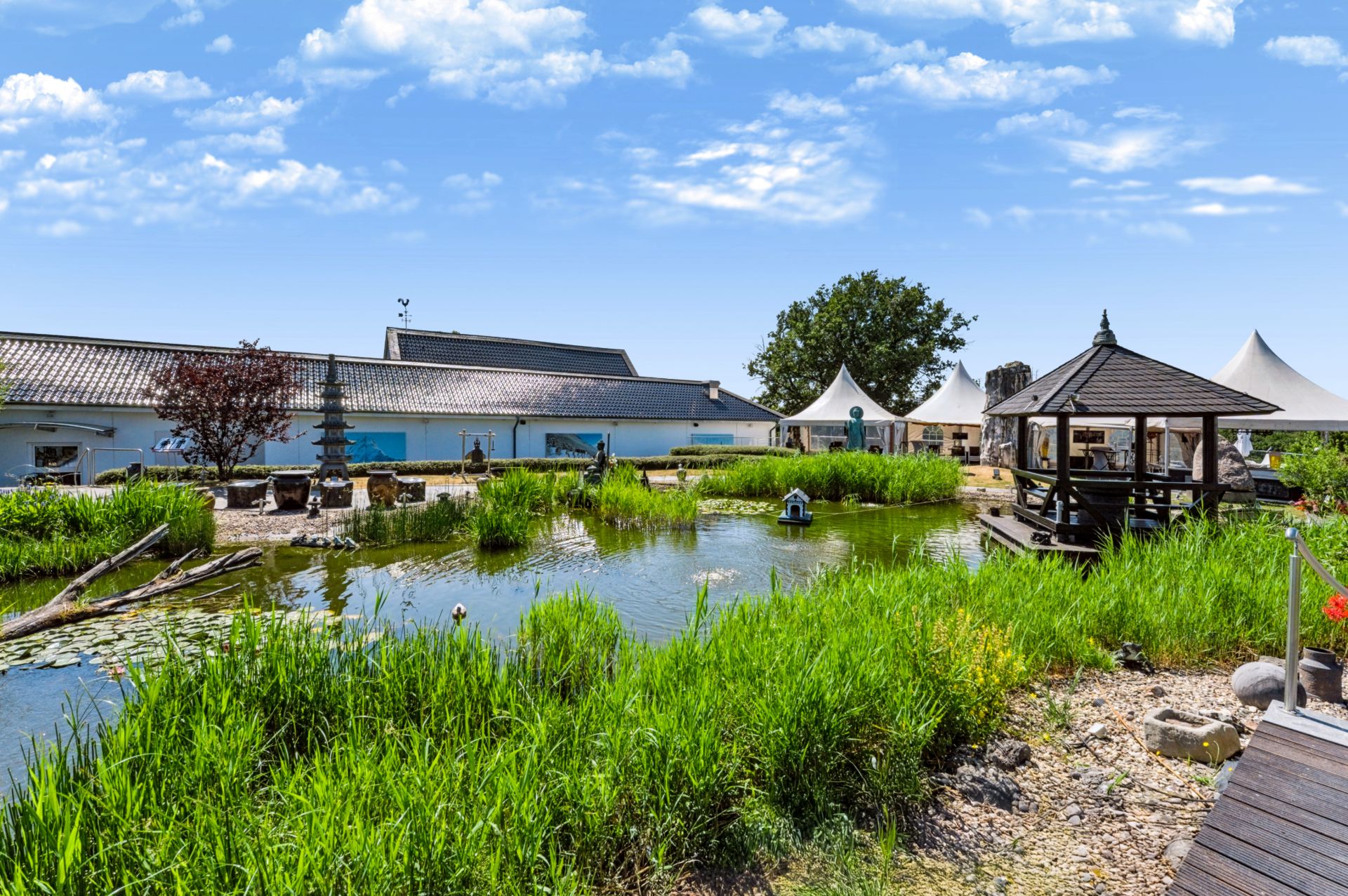 Großer Garten mit Teich, Holzpavillon und viel Grün, im Hintergrund ein langes Haus und weiße Zelte.