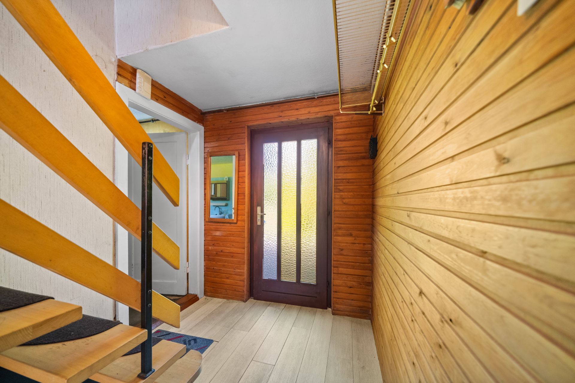 Flur mit Holzwänden, einer Glastür, Treppe und heller Holzboden.