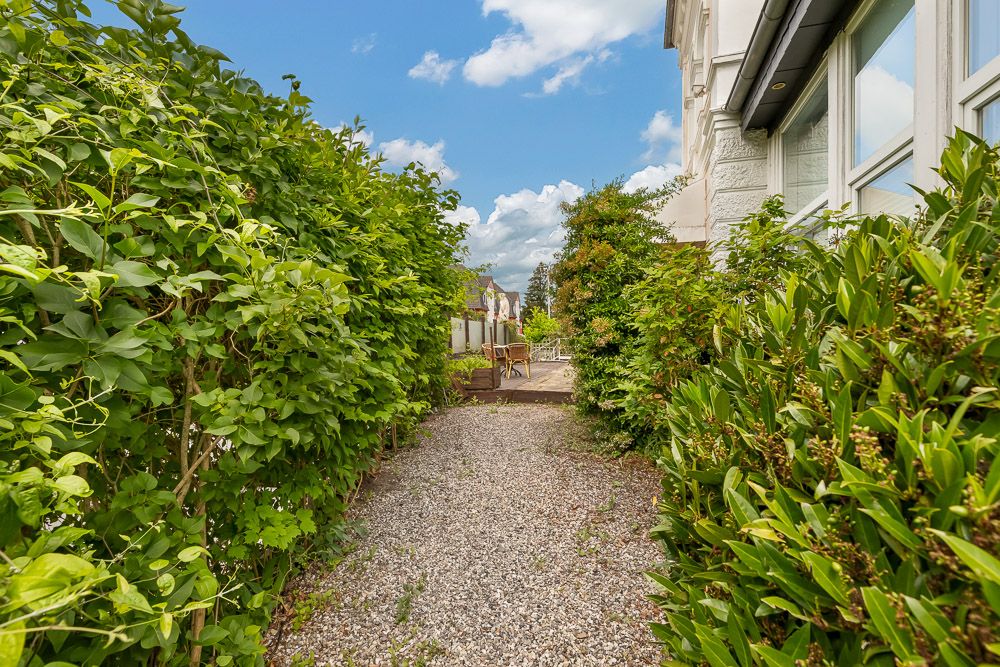 Kiesweg mit grünen Sträuchern neben einem Wohnhaus, im Hintergrund eine Terrasse mit Stühlen.