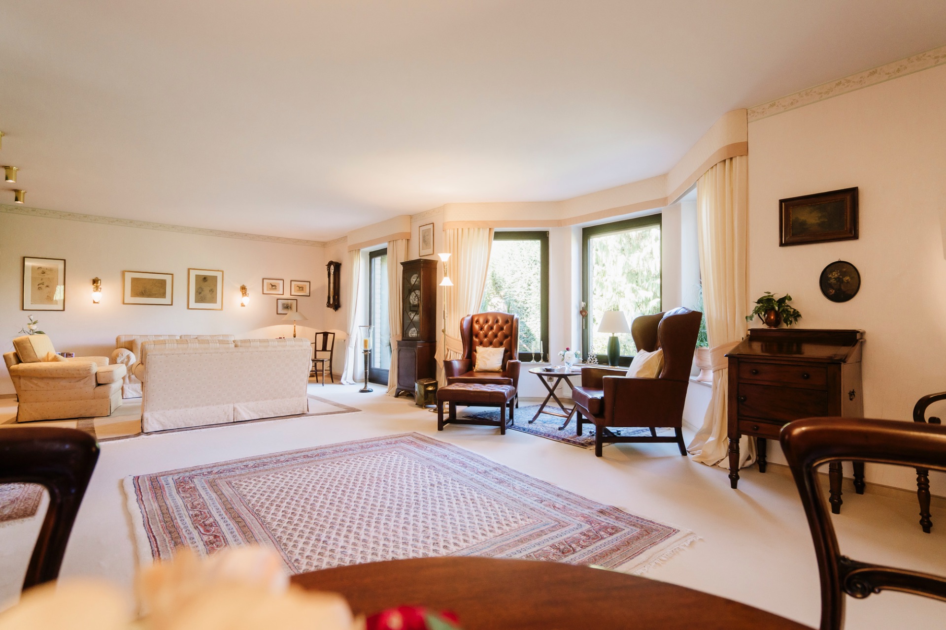 Geräumiges Wohnzimmer mit großen Fenstern, Teppich, Sesseln, Sofa und Holzmöbeln.