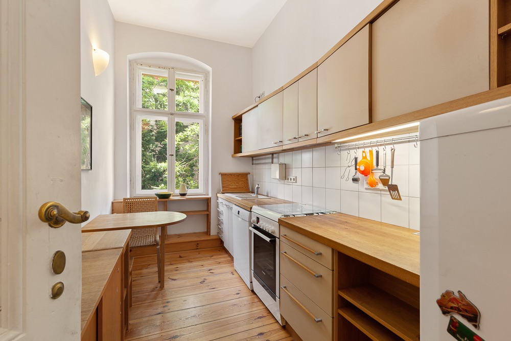 Helle Küche mit Holzfußboden, Einbauküche und kleinem Esstisch vor einem großen Fenster.