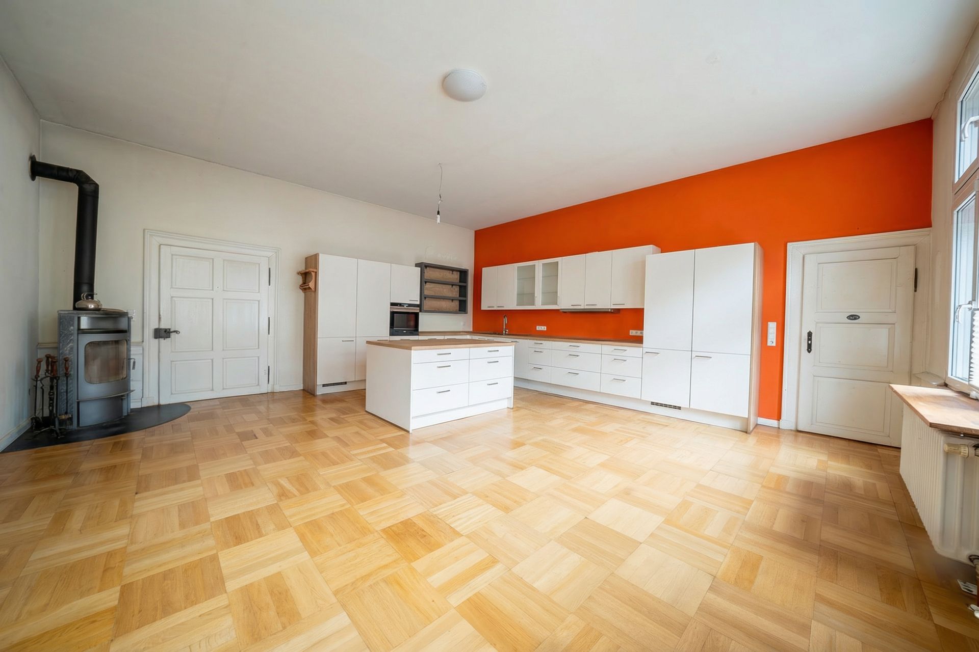 Moderne Küche mit weißen Schränken, Kücheninsel und oranger Wand, Holzofen links.