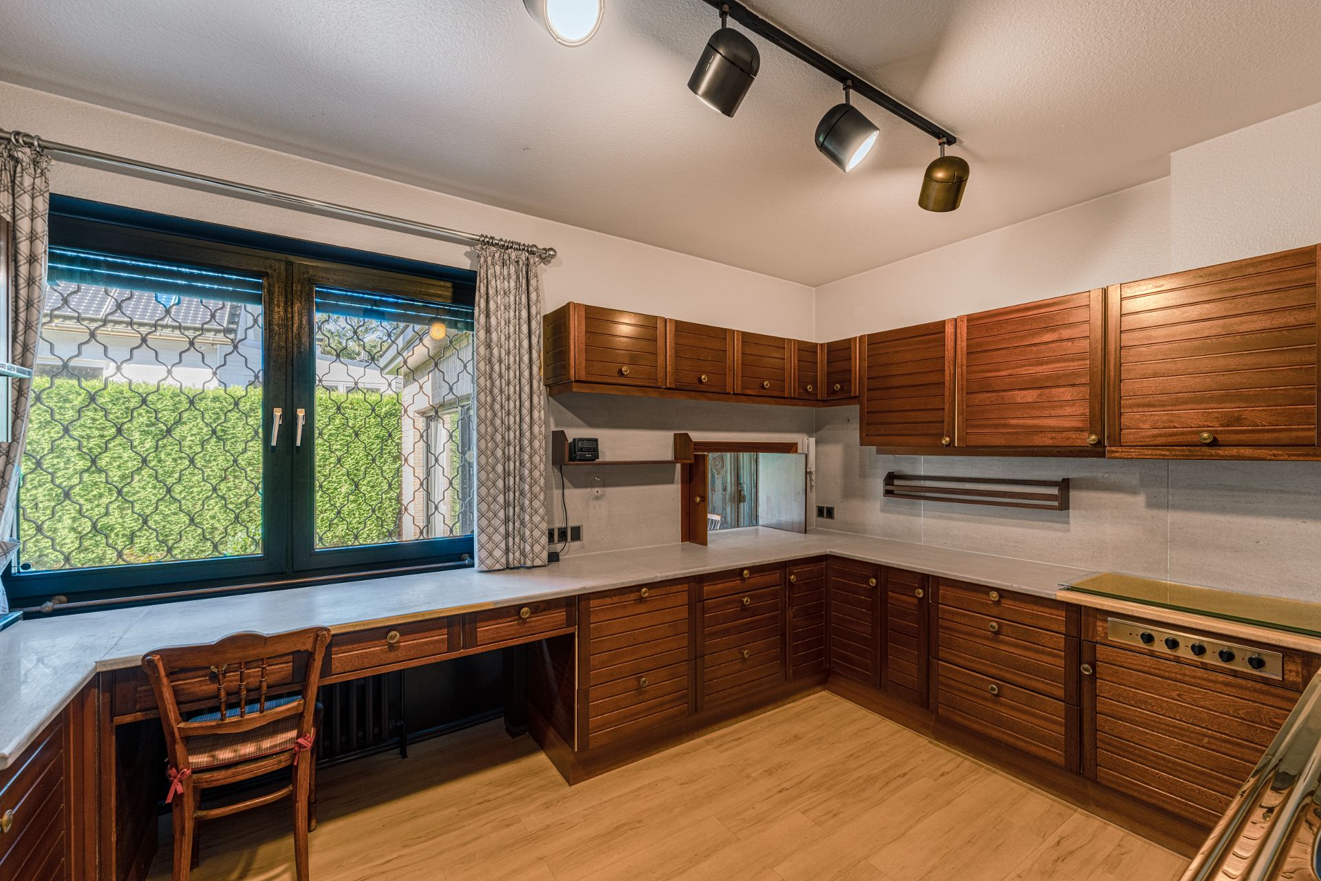 Küche mit Holzfronten, großem Fenster mit Gitter, hellem Boden und Spotbeleuchtung.