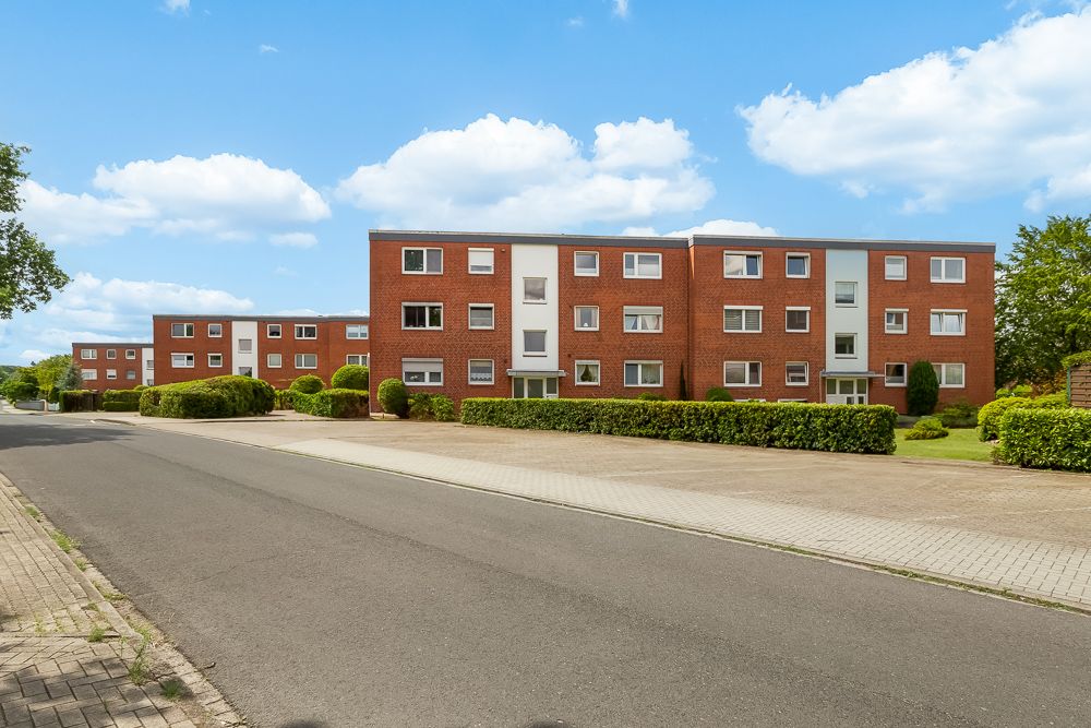 Mehrfamilienhaus mit roter Klinkerfassade, drei Stockwerken und gepflegter Begrünung davor.